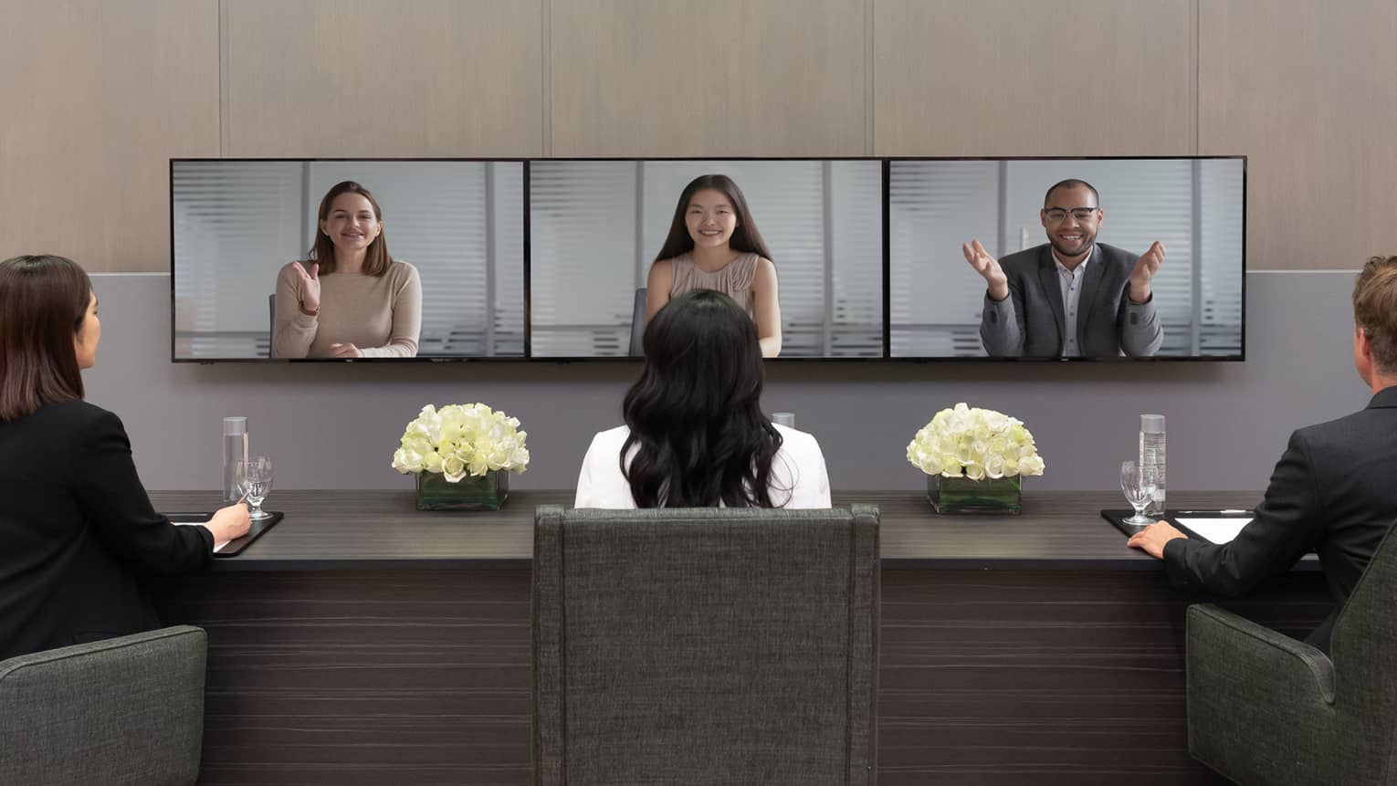 Several people in business clothes sit in front of a series of screens in a virtual meeting.