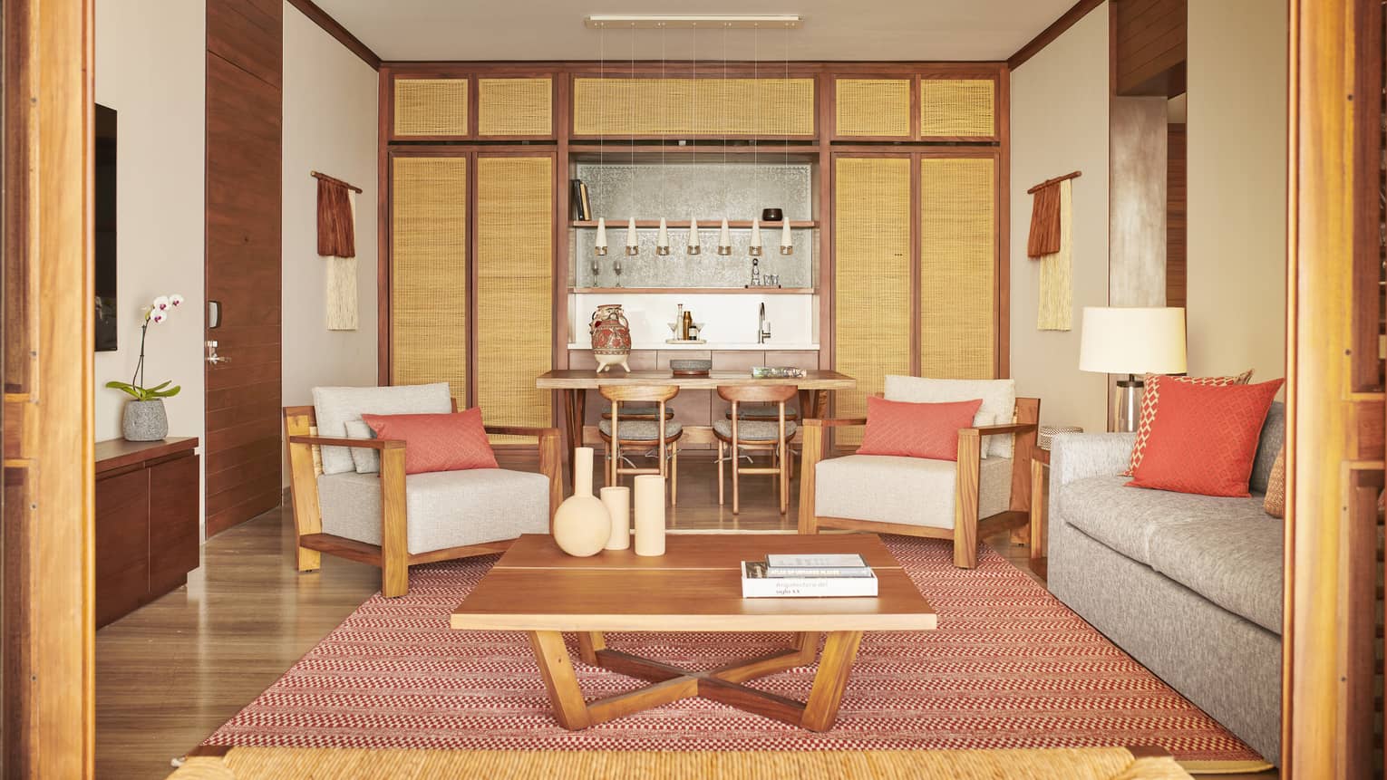A warm living room with wooden furniture and orange accents, featuring a coffee table, grey sofa and armchairs. In the background, a small dining area and kitchenette are framed by wicker panels.