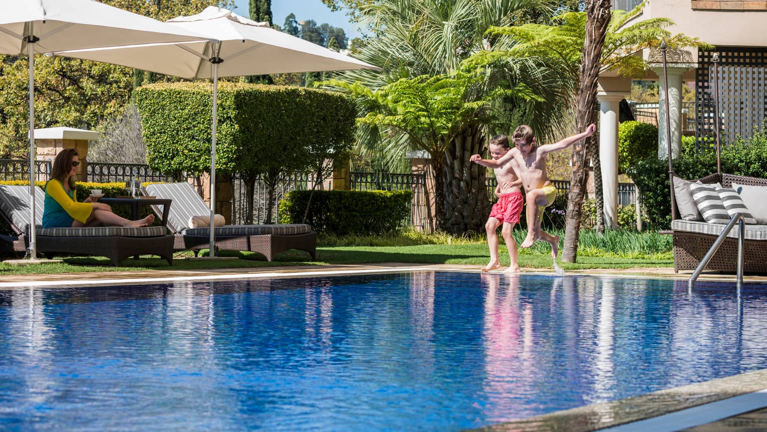 Two kids leap into outdoor swimming pool, mom watches from lounge chair