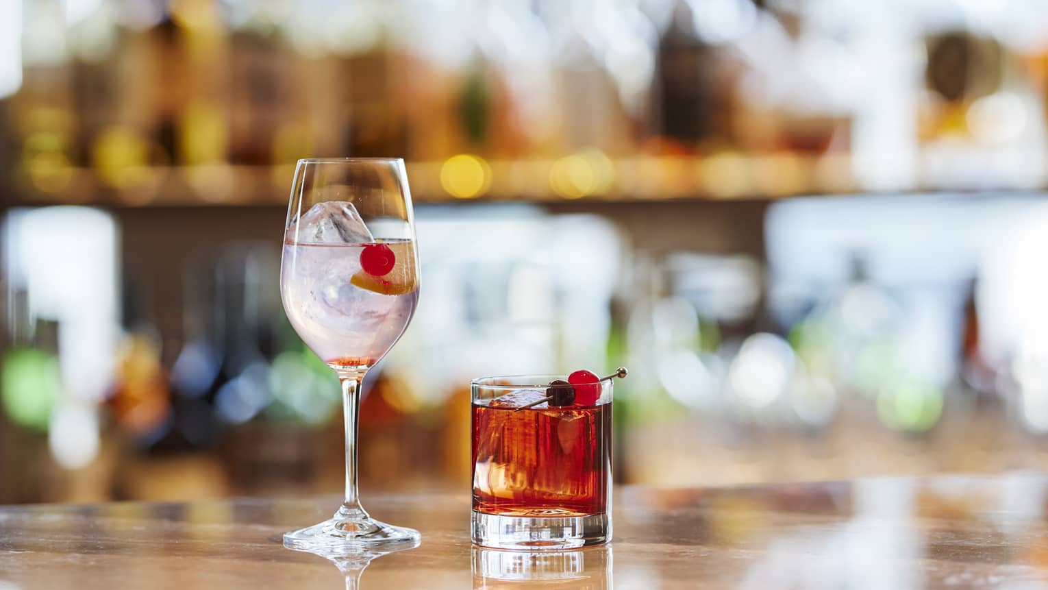 Two cocktails, one is stemmed glass, one in tumbler, with Maraschino cherry garnish on bartop