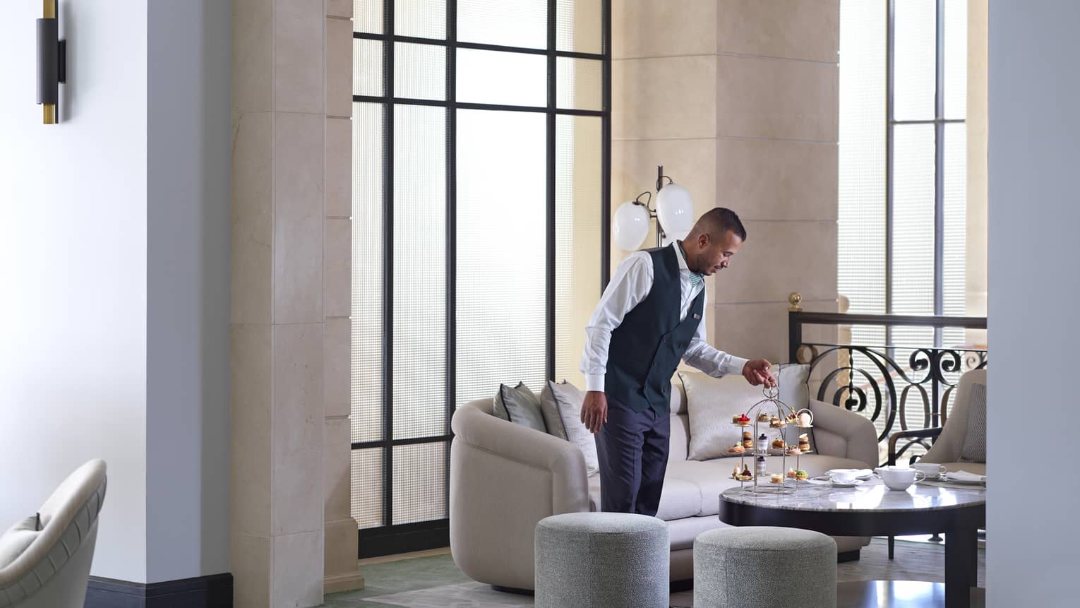 A service member in a navy waistcoat and slacks sets down a three-tiered tray of treats for Afternoon Tea in the Foyer Lounge. 