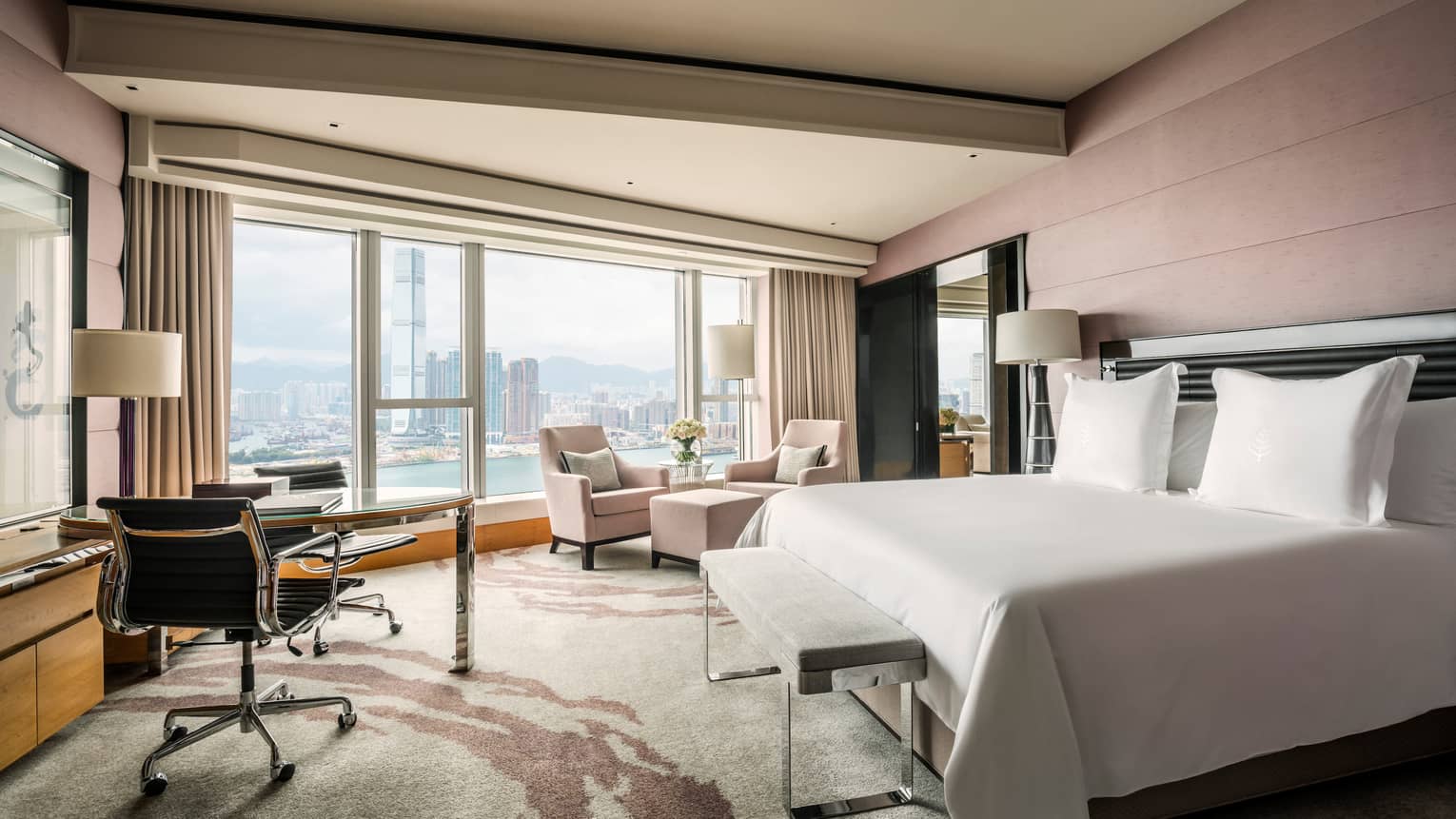 Hong Kong hotel room bed, desk, two armchairs and ottoman in front of floor-to-ceiling window, waterfront view