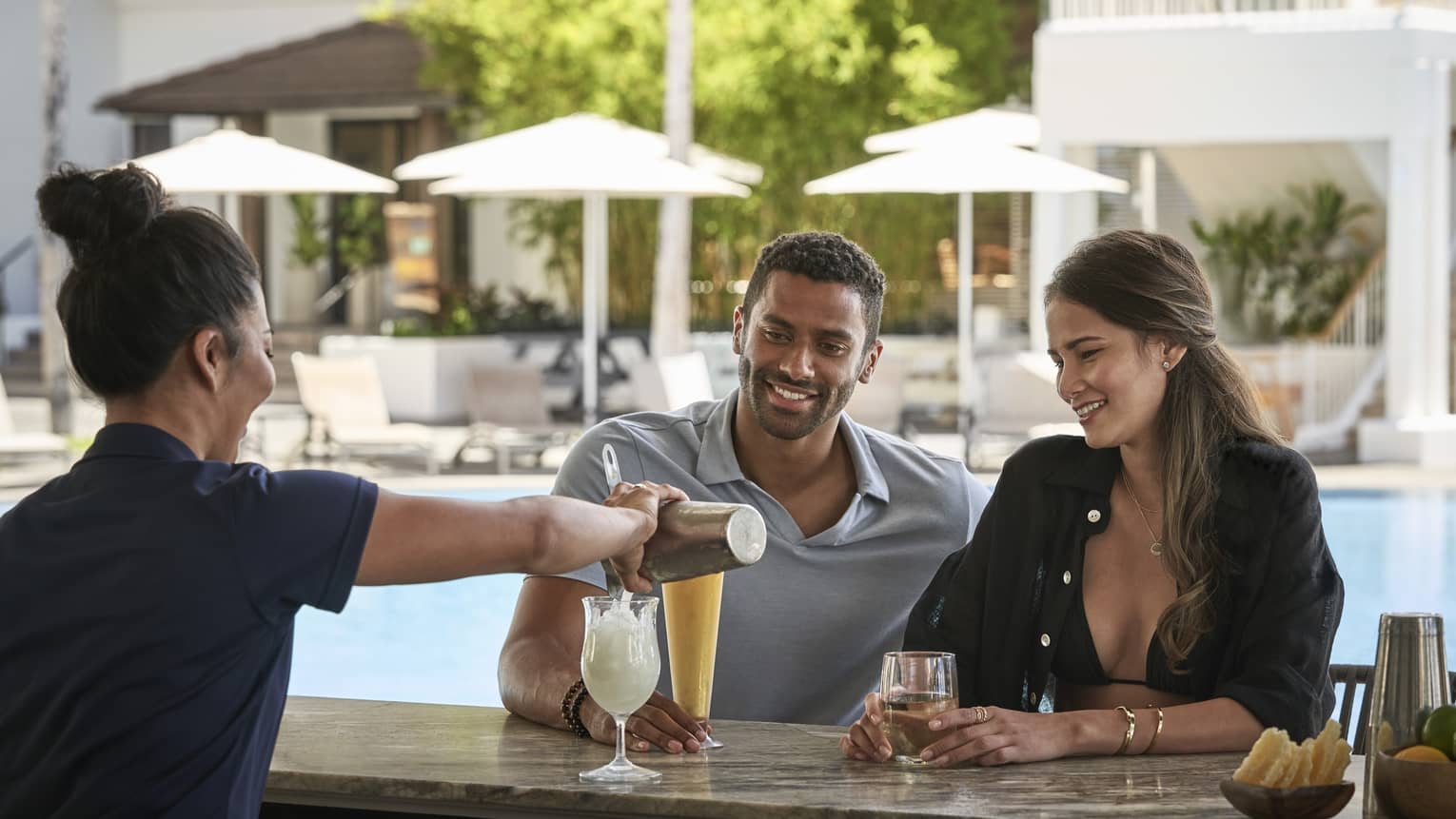 Female bartender pours a tropical drink for man and woman at poolside bar