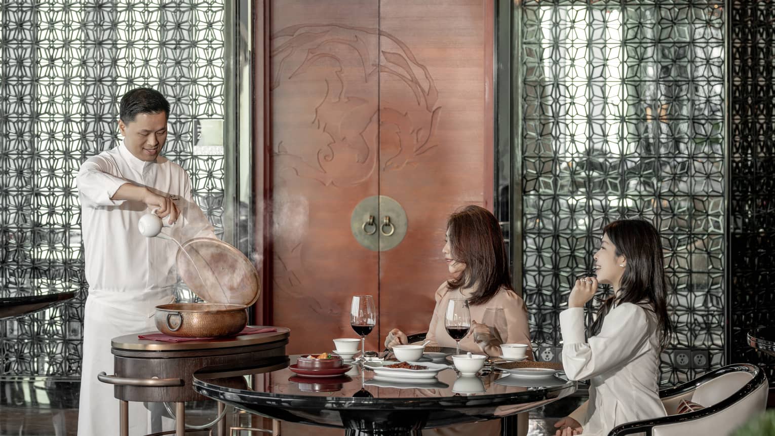 Two female diners seated at table watch as chef pours liquid into large copper pot 