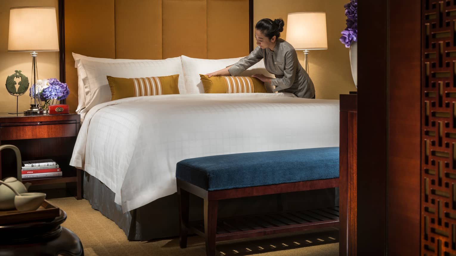 Hotel staff makes bed, adjusts gold accent pillow on white pillows, linens