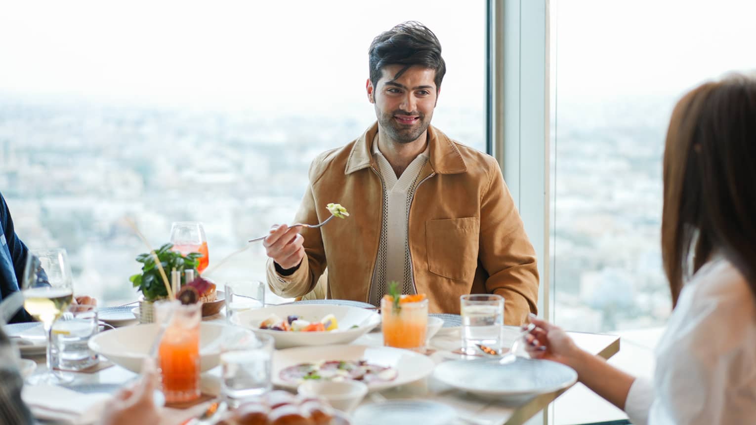 Man sits at restaurant table with a wall of windows behind him overlooking a city skyline