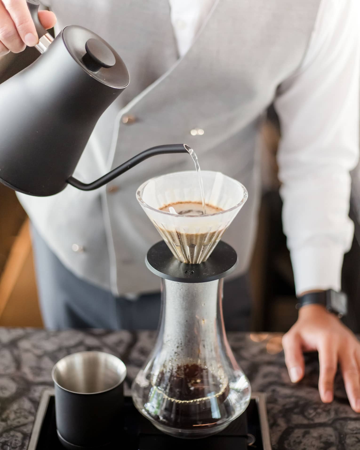 Barista holds black teapot, pours hot water over coffee in cone filter and glass jug on tray