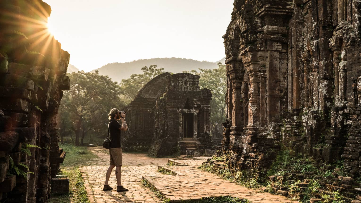A tourist photographing ancient stone ruins, surrounded by lush greenery and misty hills in the background.