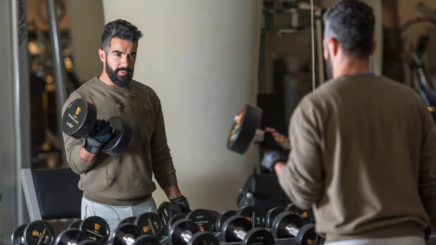 Man with beard wearing casual sweatshirt and sweatpants stands looks in gym mirror, lifts large hand weights