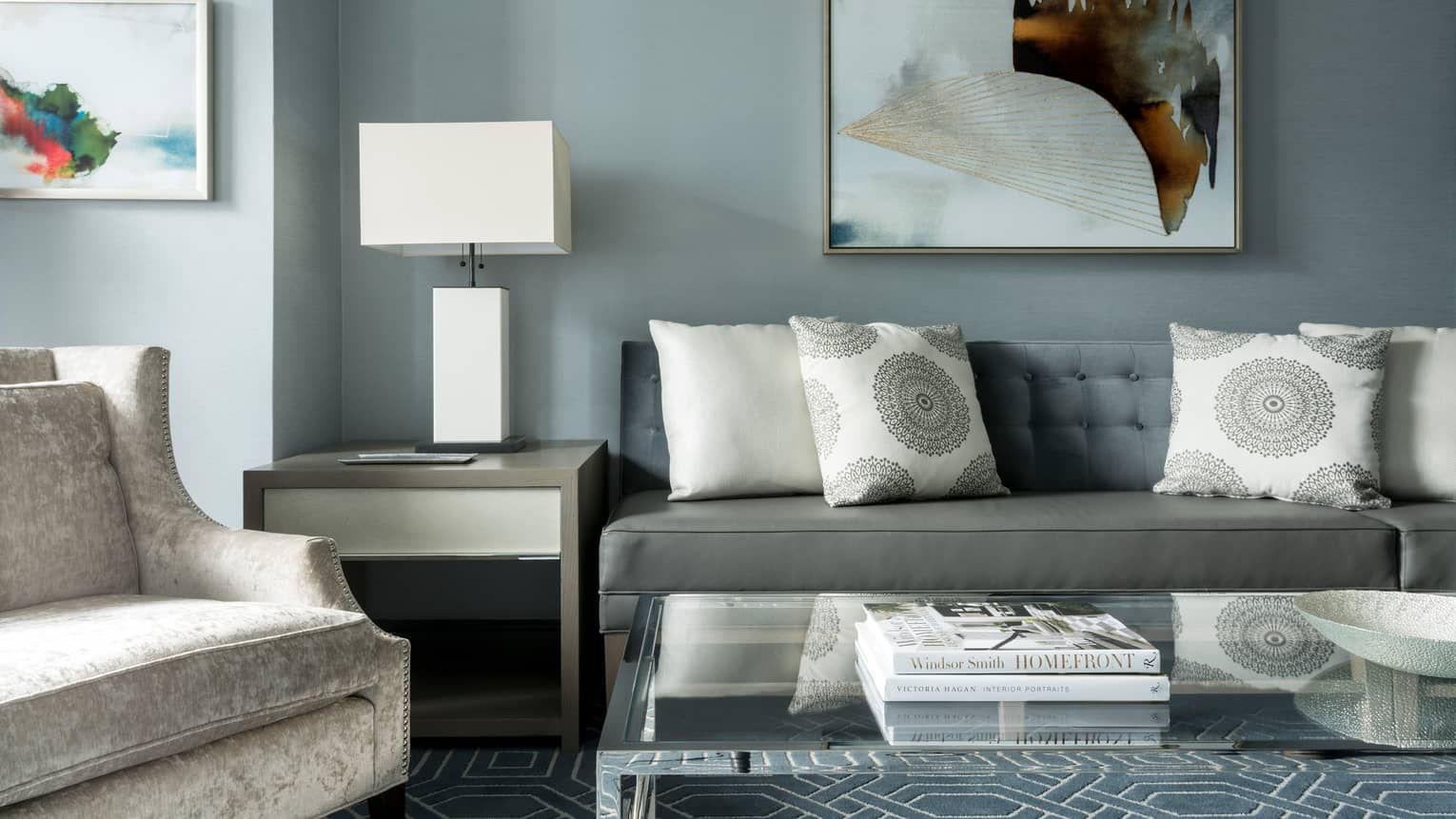 Midtown Suite long grey sofa with white accent pillows below abstract painting, table, lamp, armchair