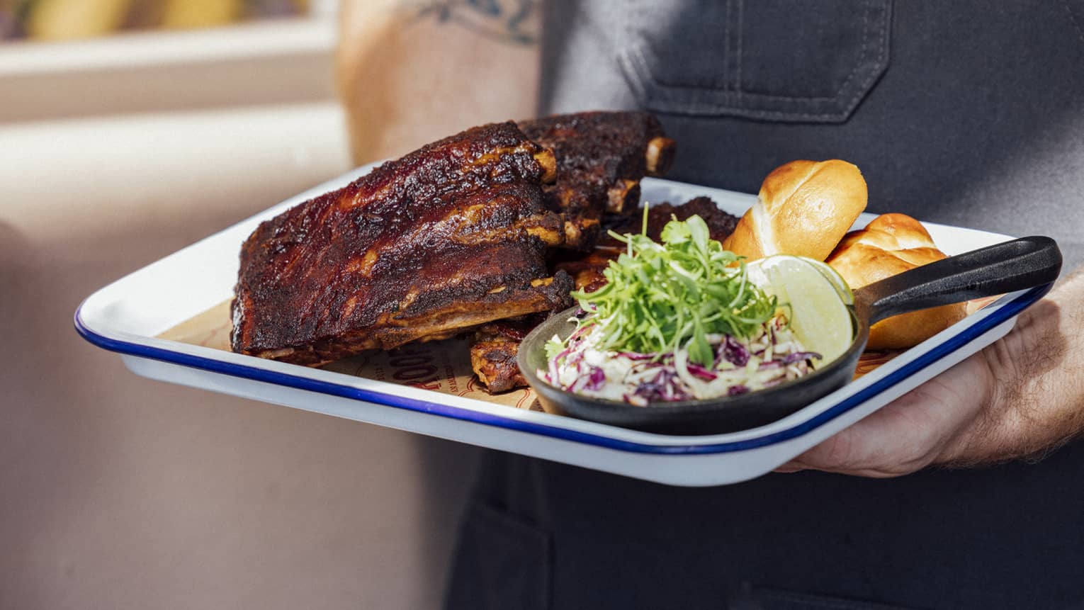 A person holds a rectangular plate with a meal of cooked meat and sides.