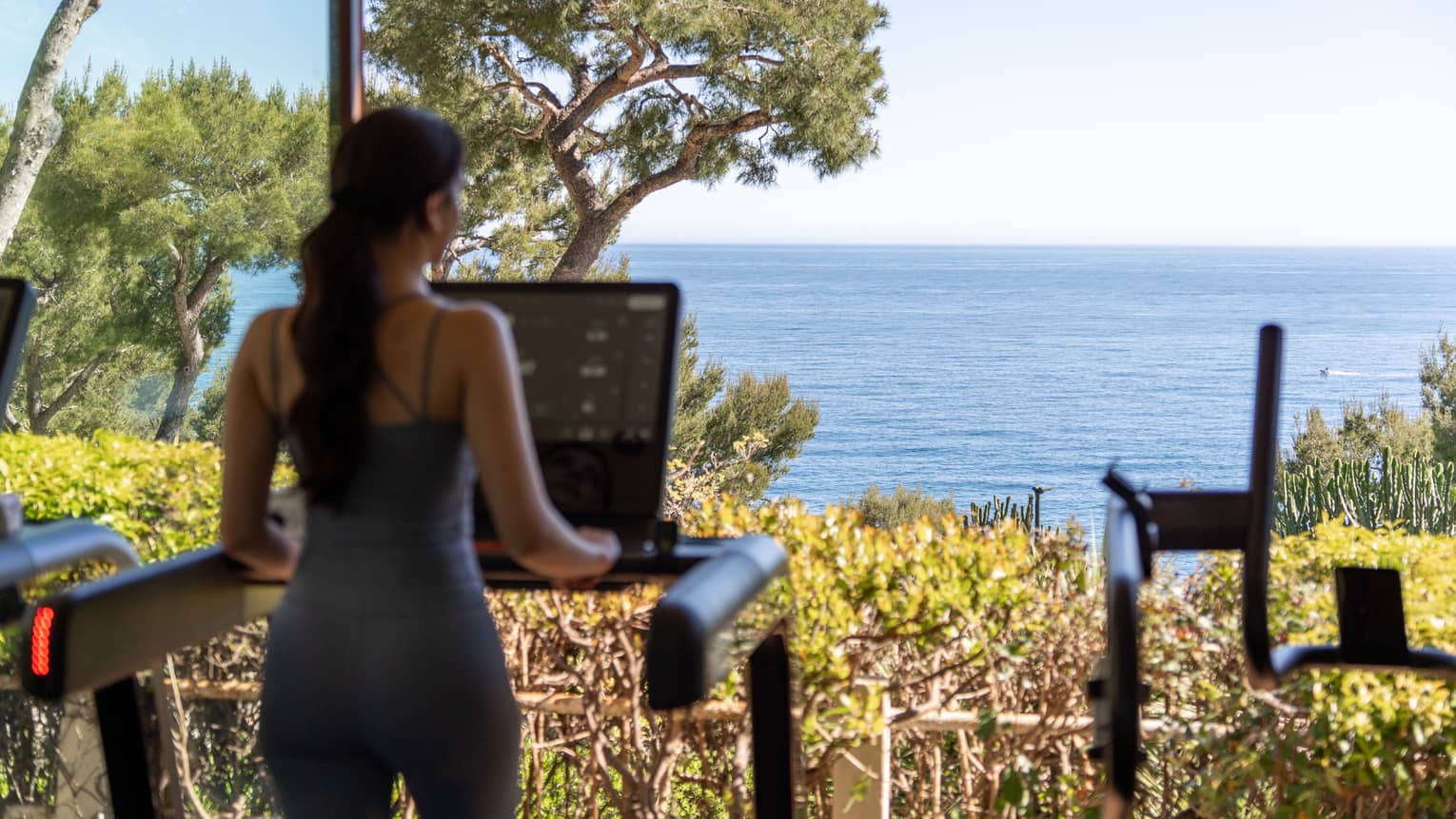 A person stands on a treadmill facing large windows with landscape and ocean views.
