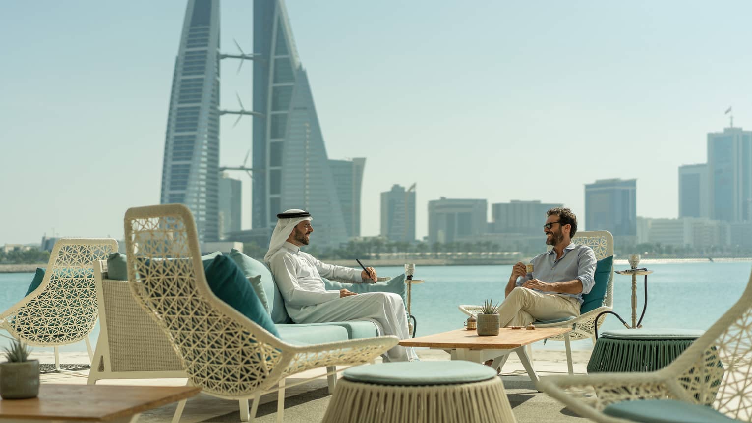 Two men sit in lounge chairs at rooftop Shisha Lounge overlooking sunny skyline, water