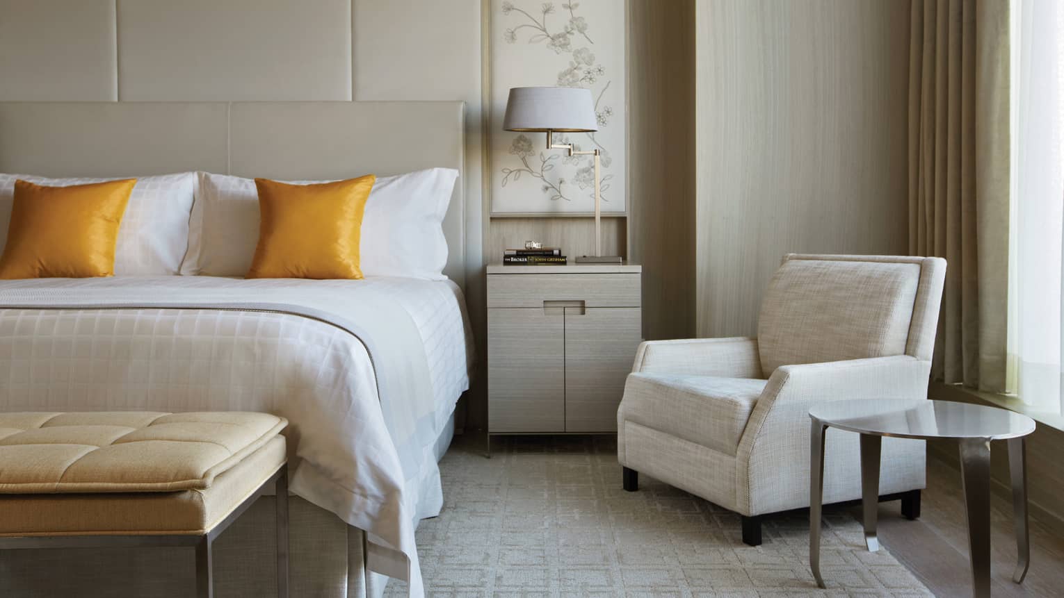 Hotel suite bed with golden yellow pillows, plush bench by nightstand, grey armchair, window