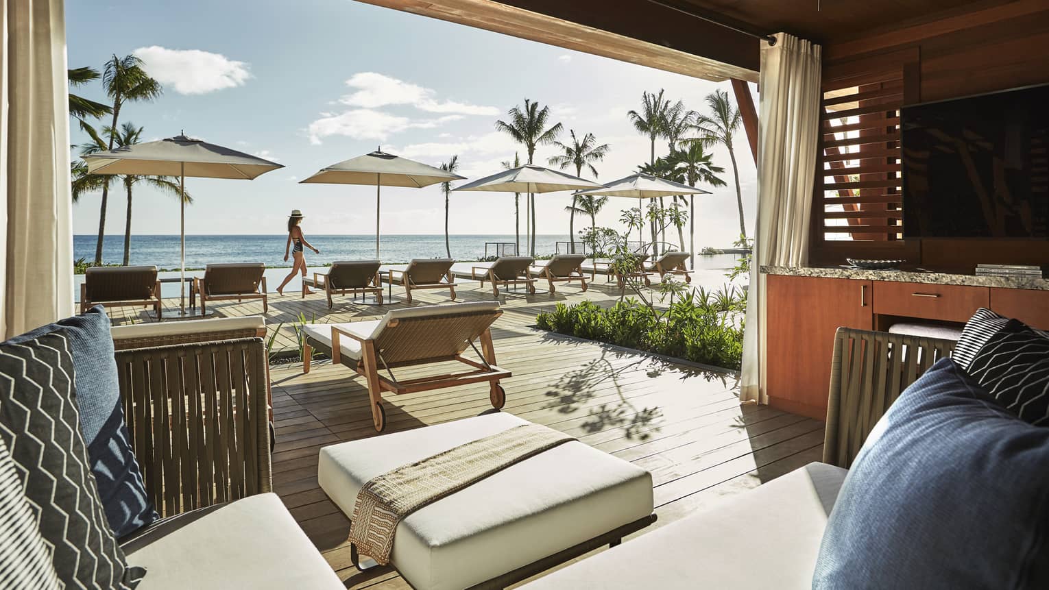 Plush patio lounge chairs under wood cabana, woman walks by pool deck