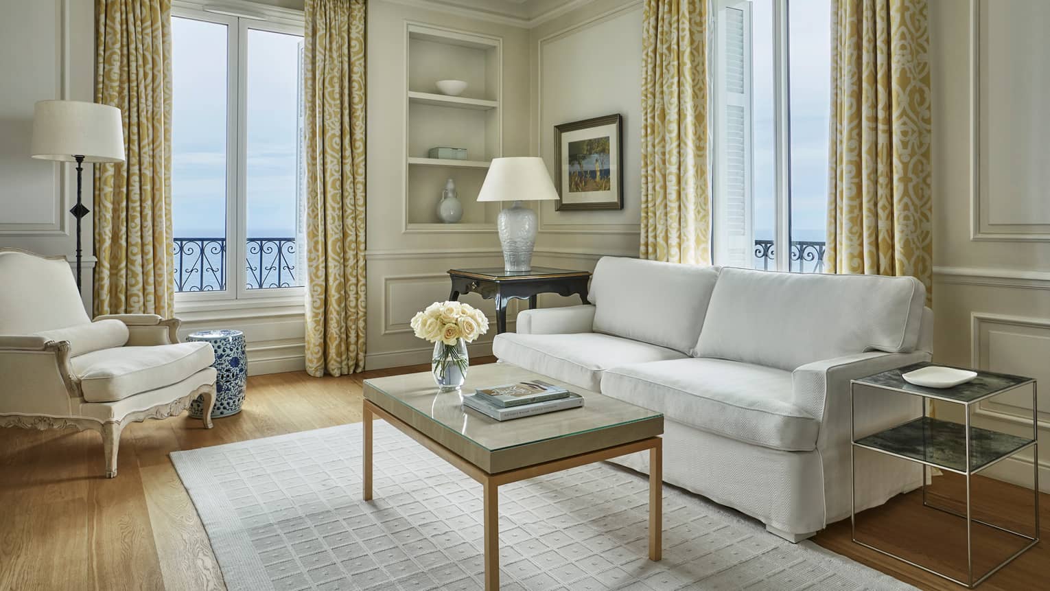 Modern white sofa, armchair, rug, table with books, flowers, corner windows