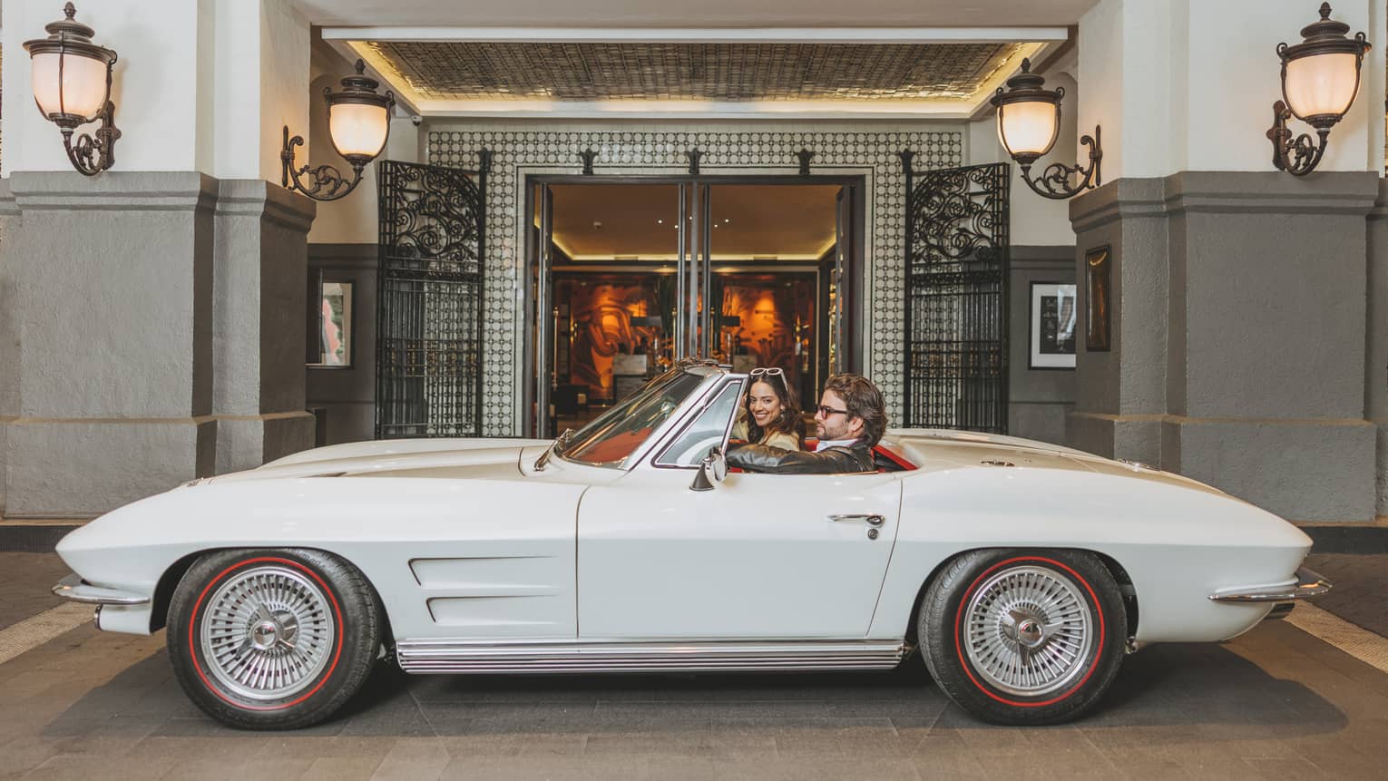 A couple sitting in a vintage white convertible parked outside a luxury hotel entrance, surrounded by ornate lighting and elegant decor.