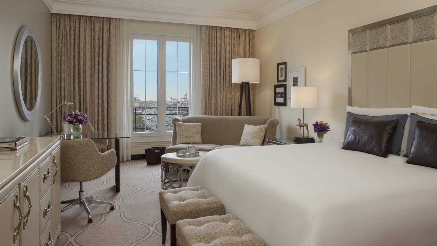 City-View Room with clean white decor, bed with tall padded headboard with silver details, brown pillows, plush benches, chairs