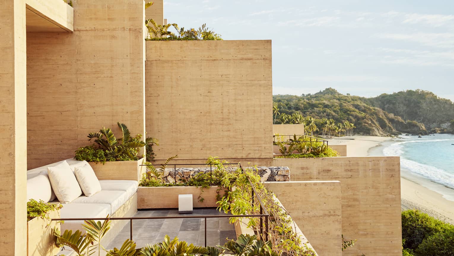 A beachfront terrace with modern concrete walls, lush greenery and a built-in seating area. The view overlooks a serene beach with palm trees and distant hills.