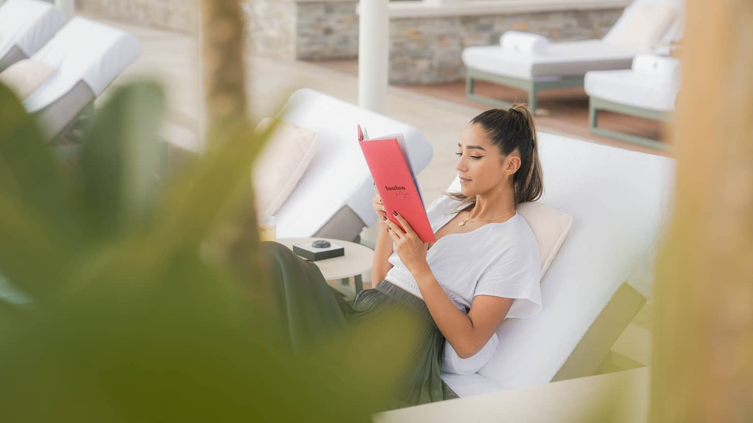 Person wearing white t-shirt and dark-grey skirt reclines on a poolside lounge chair reading a red book