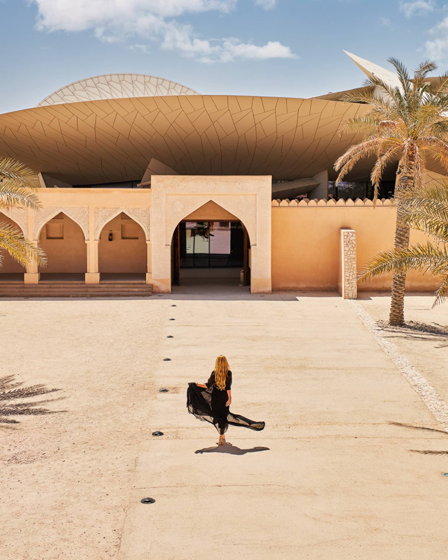 Person wearing long black flowing dress walks along a sandy walkway leading up to an Arabesque-style building