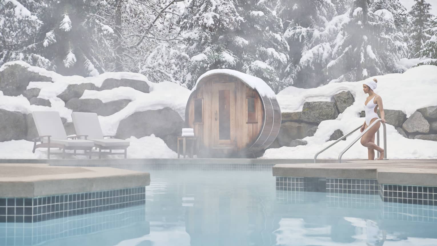 A woman in a white swimsuit enters the outdoor heated pool as steam rises around her. She is surrounded by snow-capped trees.