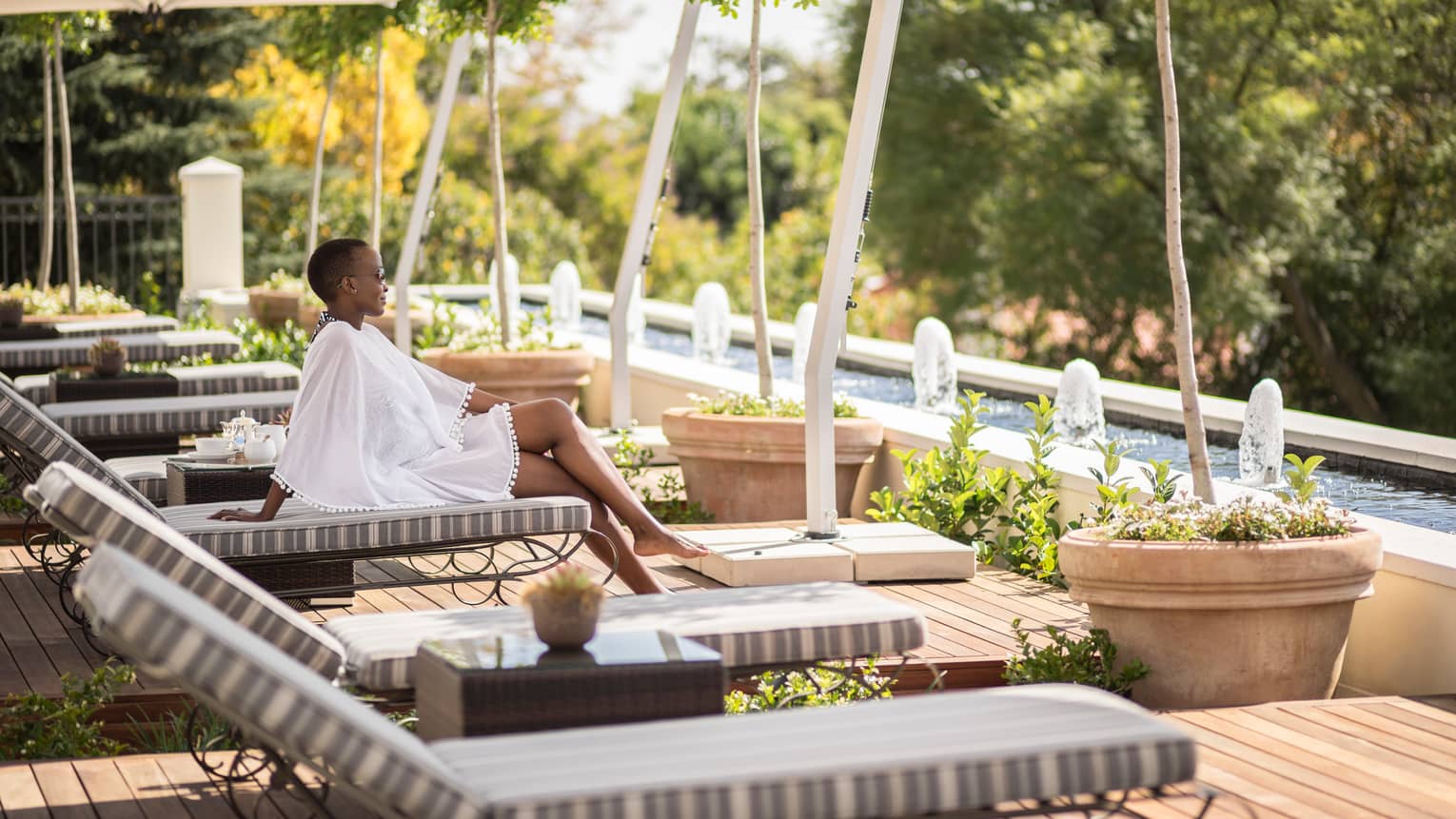 Woman in flowing white dress sits on lounge chair in row near outdoor swimming pool