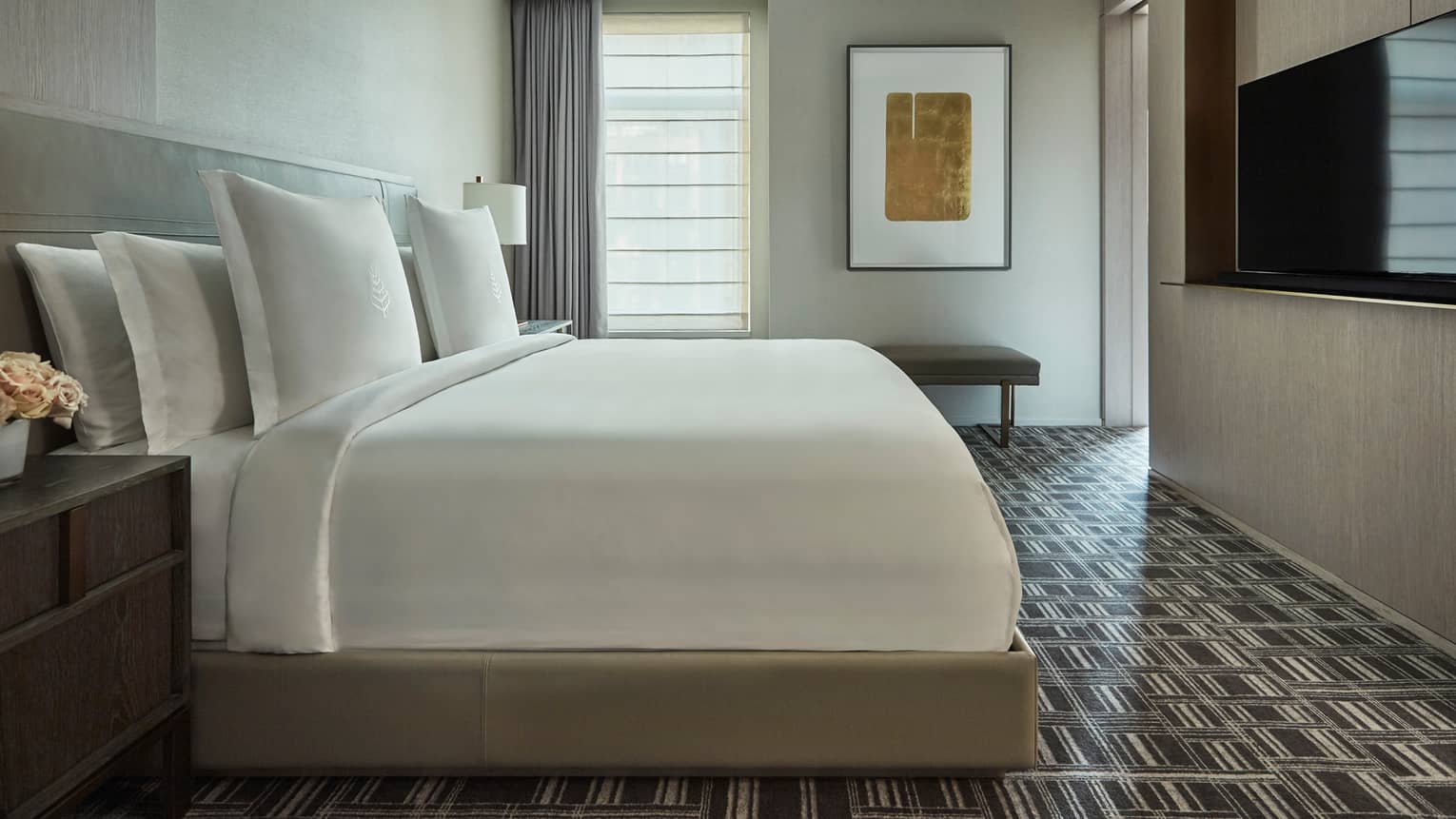 Hotel bedroom with a large white bed, nightstand, patterned rug, wall-mounted TV, wall art and a bench near the window with sheer blinds