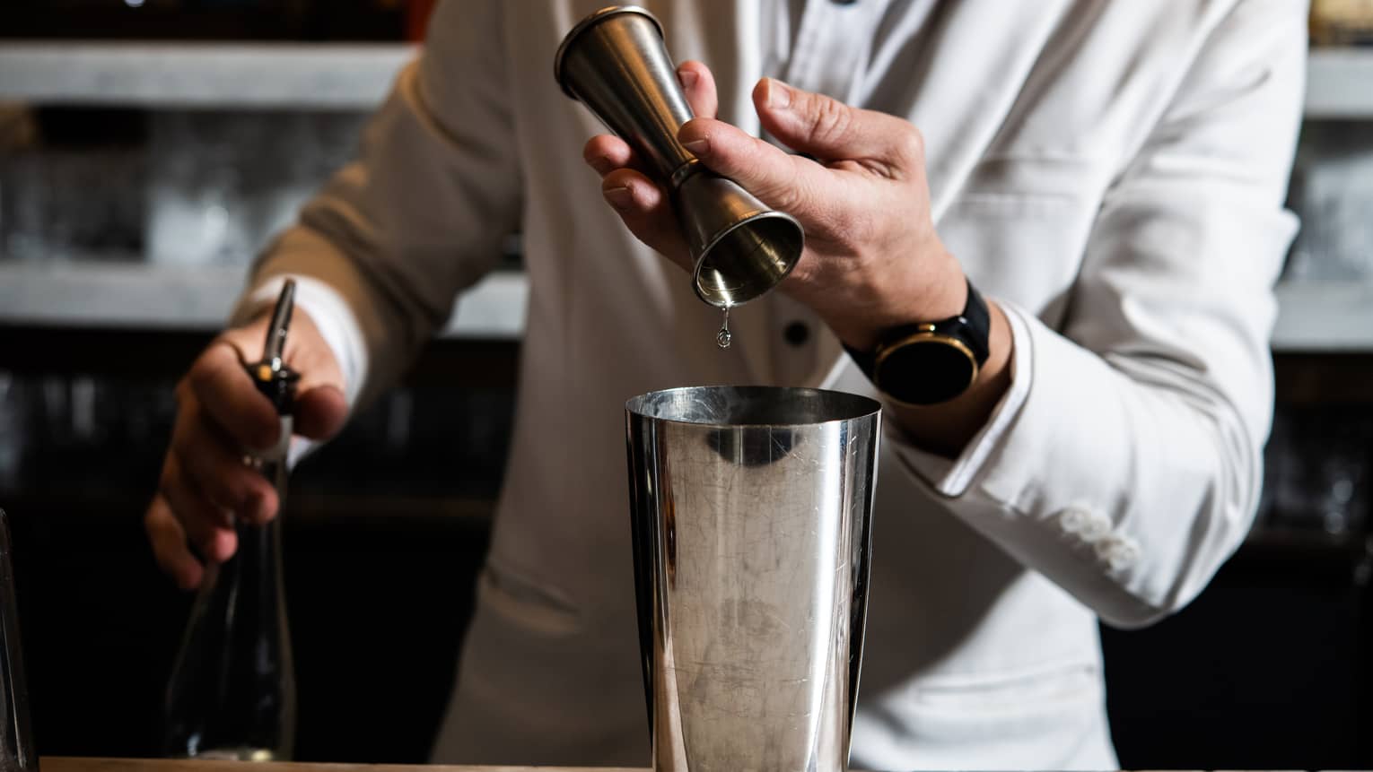 A man in an all-white suit mixes a cocktail in a silver shaker.