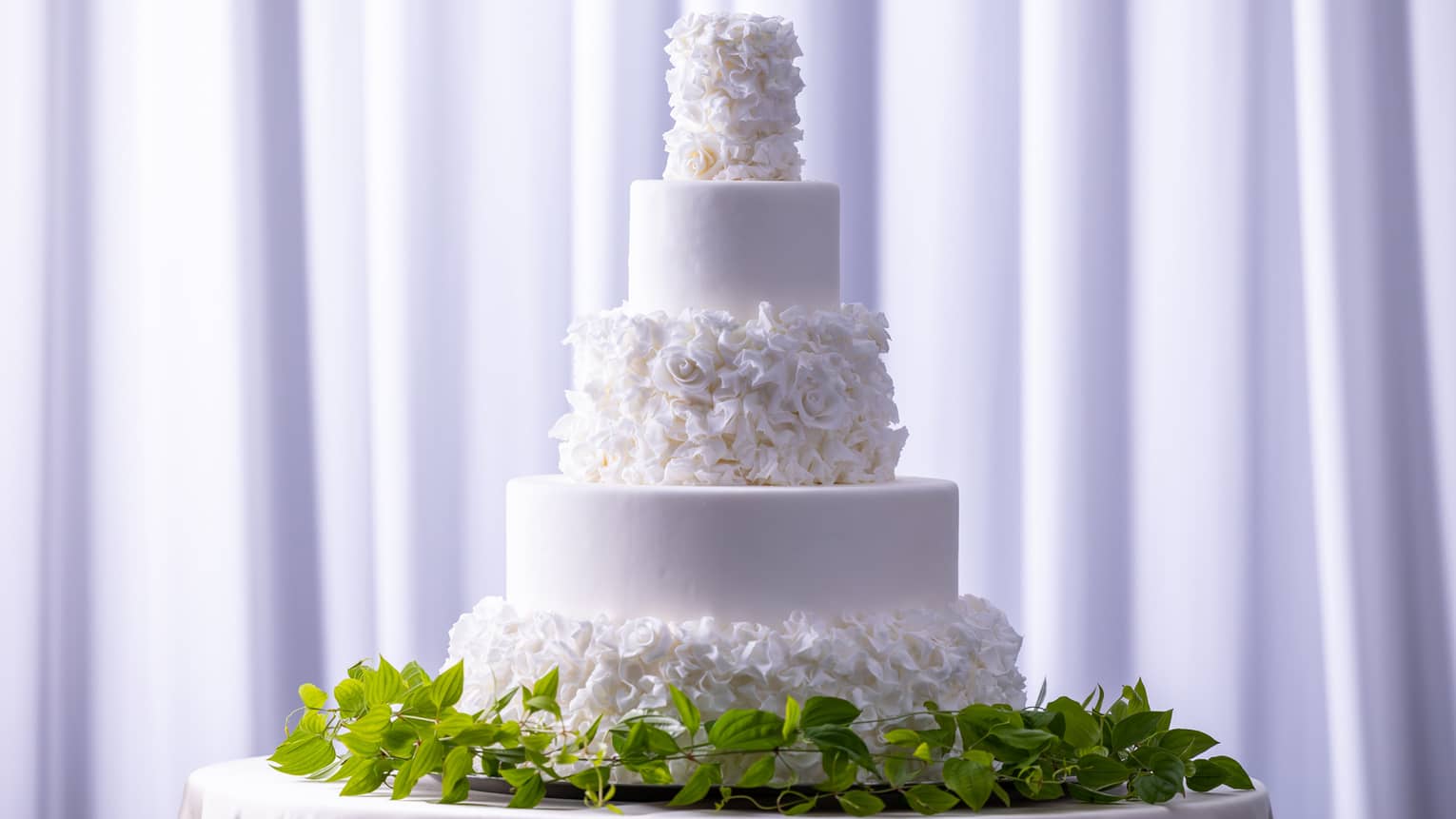 A white floral wedding cake on a stand surrounded by green vines