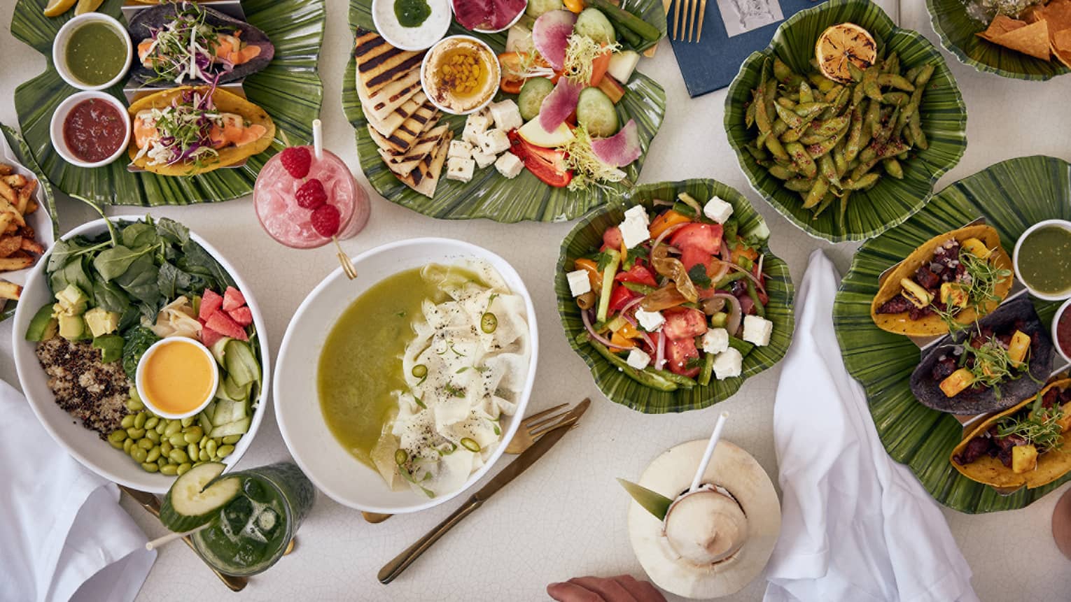 Assortment of vibrant dishes including salads, tacos, edamame, soup and tropical drinks served on a table with banana leaf platters