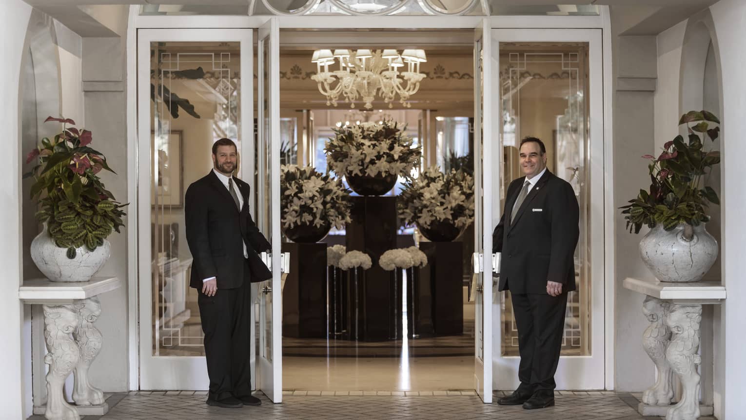 Doormen open front entrance doors to lobby with white flower arrangements on pedestals