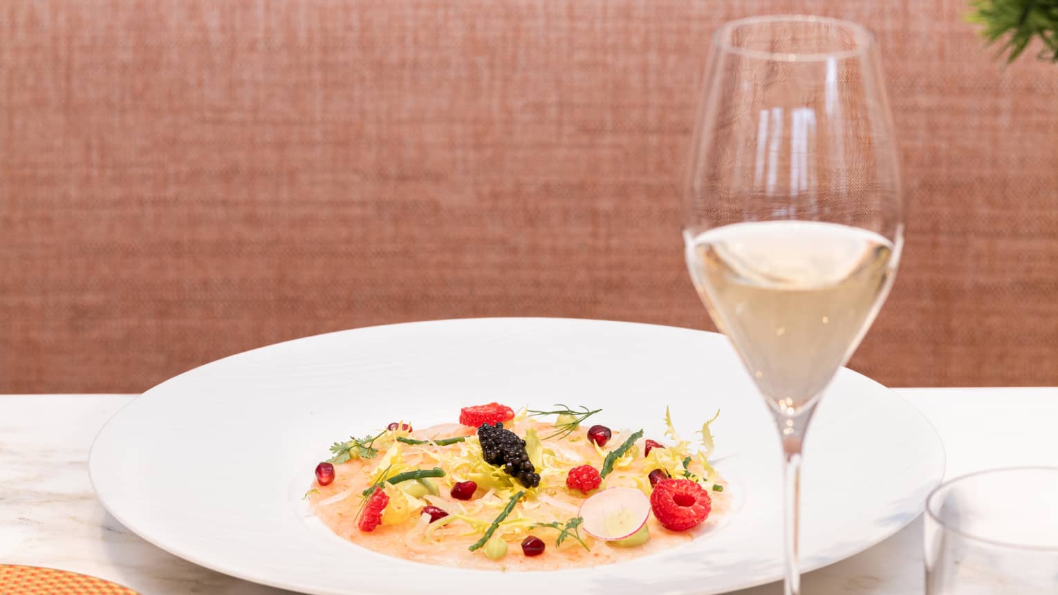 A wide bowl of saffron-coloured pasta garnished with raspberries, caviar and herbs, served with a glass of white wine.