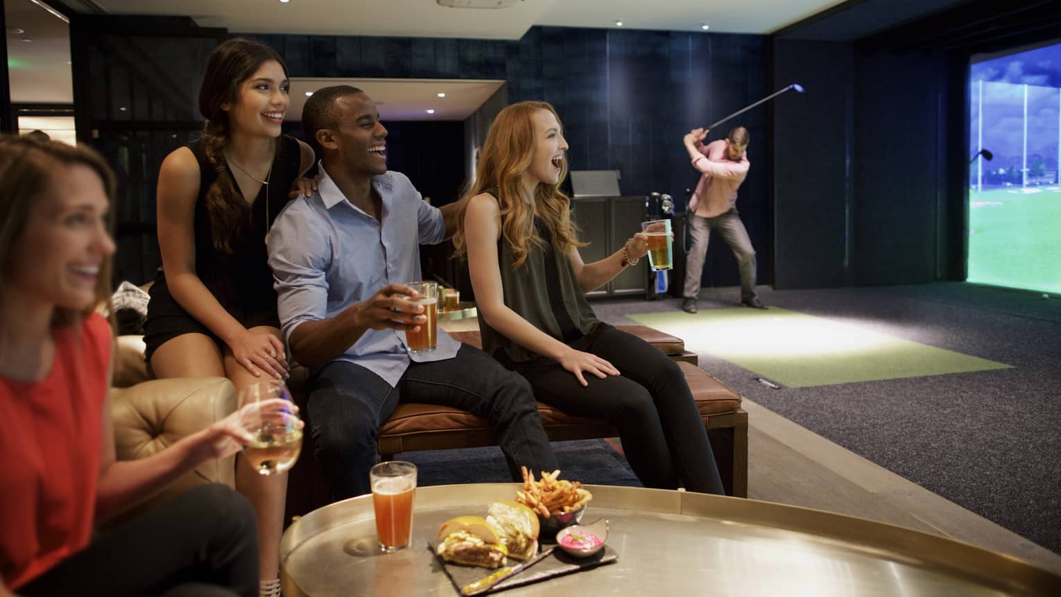 A man and three woman sip beer and cheer on a sports team in the foreground, while another man golfs in the background of a function room at four seasons hotel houston 