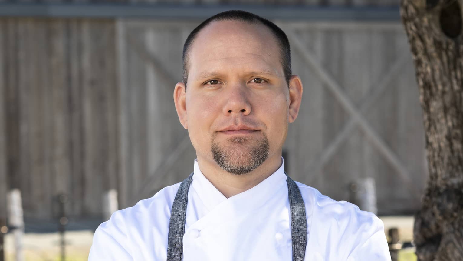 A chef standing outside near a tree and in front of a barn.
