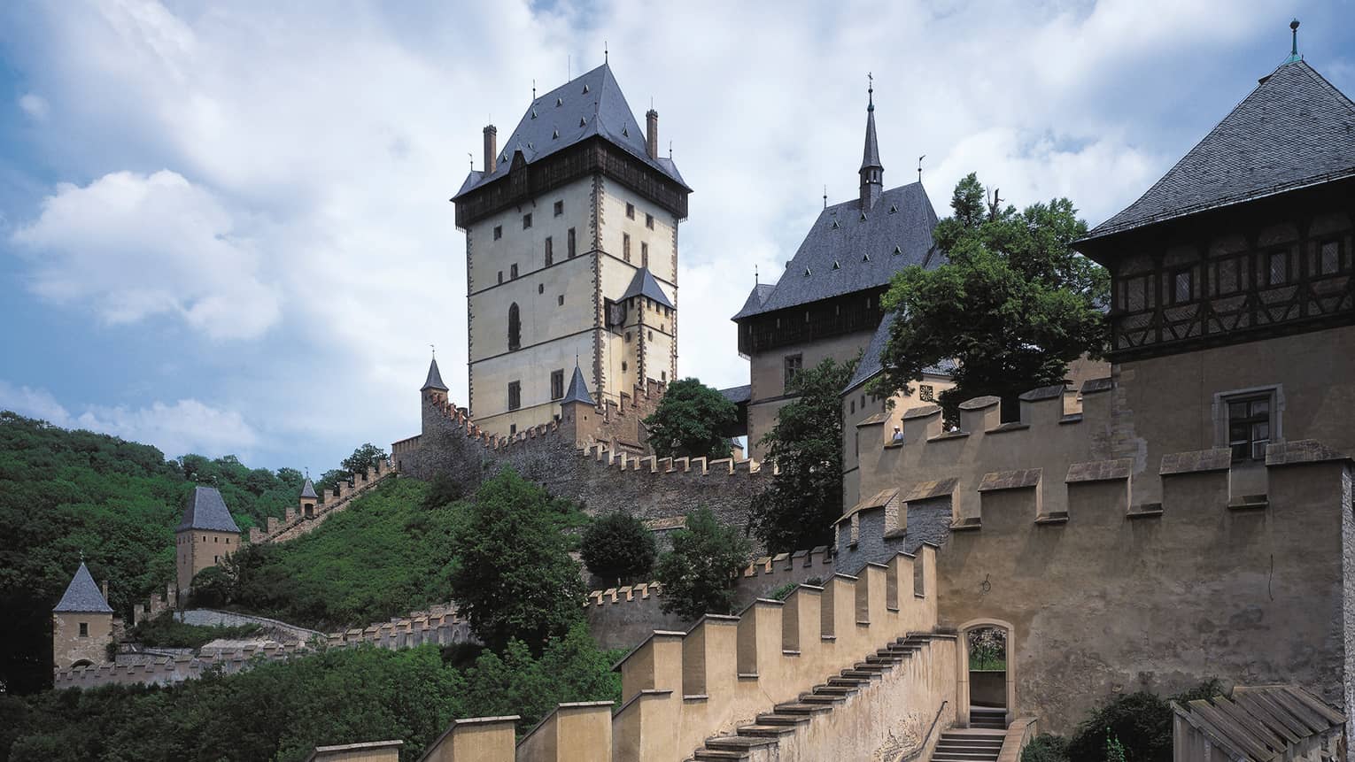 A historic castle with tall towers, sloped rooftops and fortified walls, surrounded by greenery and set against a cloudy sky.