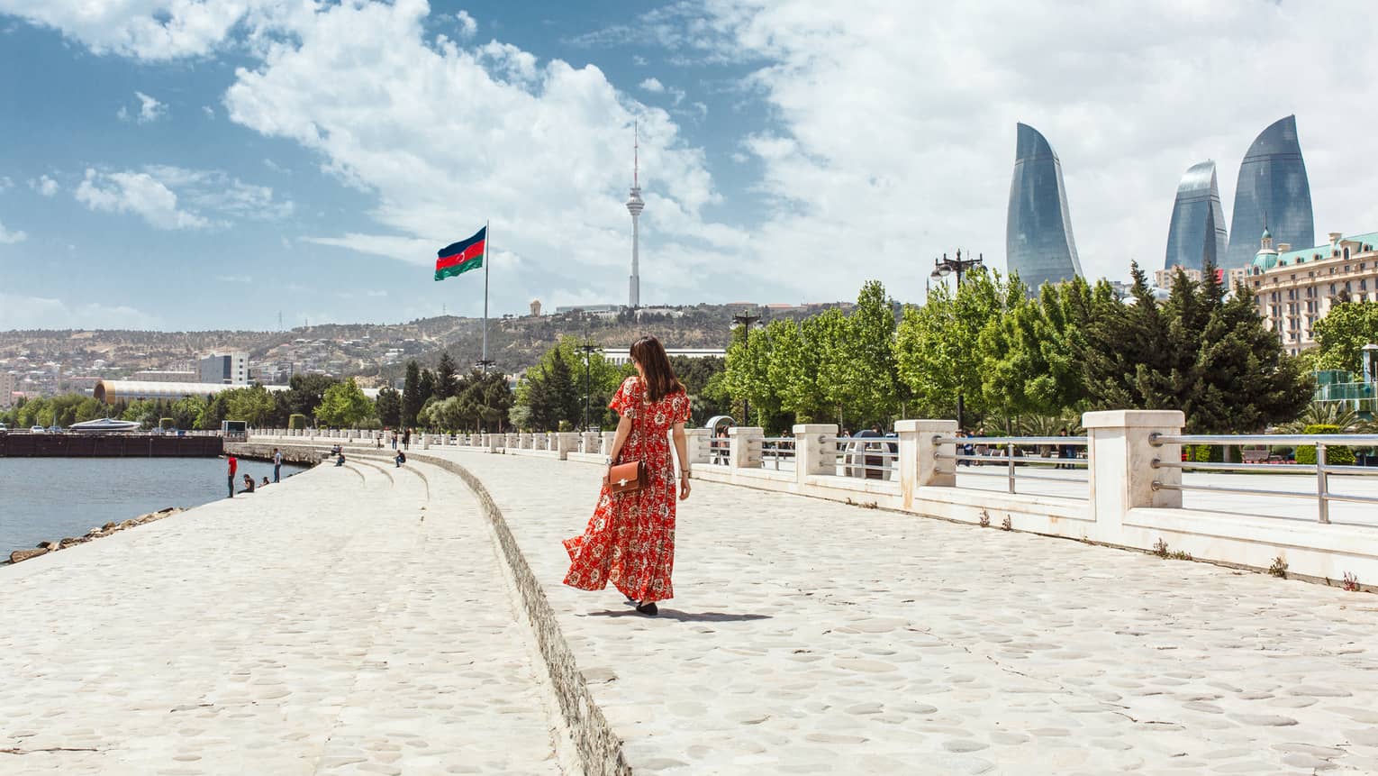 Woman wearing red dress walks along stone waterfront, skyscrapers in background