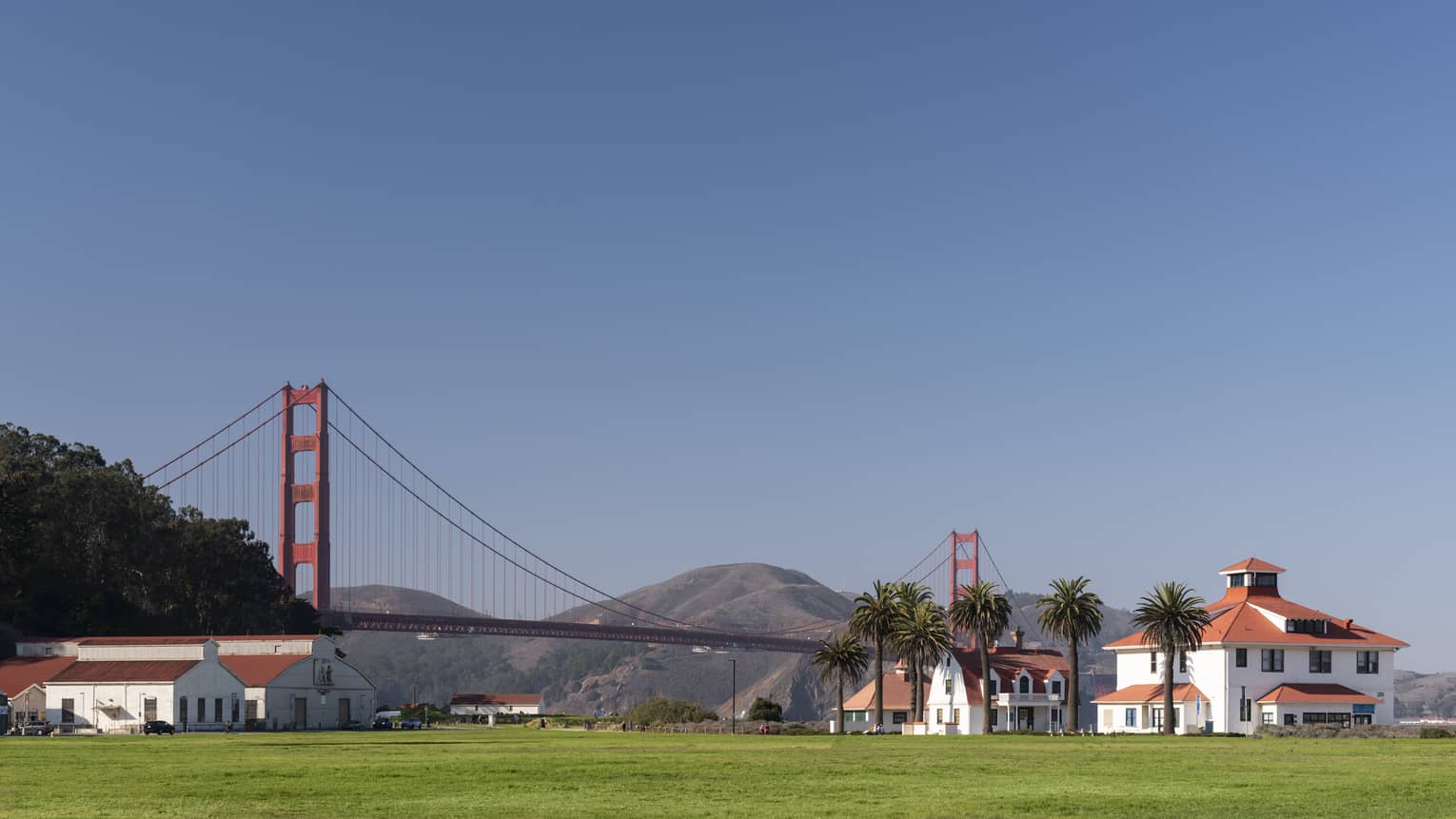 Red-roofed white buildings at the far end of a green field with the Golden Gate Bridge, rolling hills and a blue sky beyond.