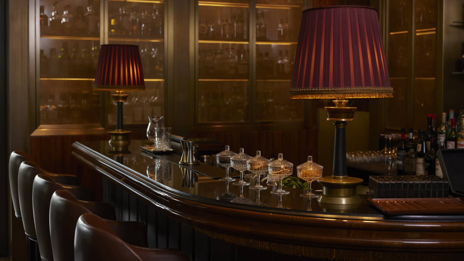 Curved dark wood bar counter with two tall red lamps, various glassware and five brown leather stools next to the counter