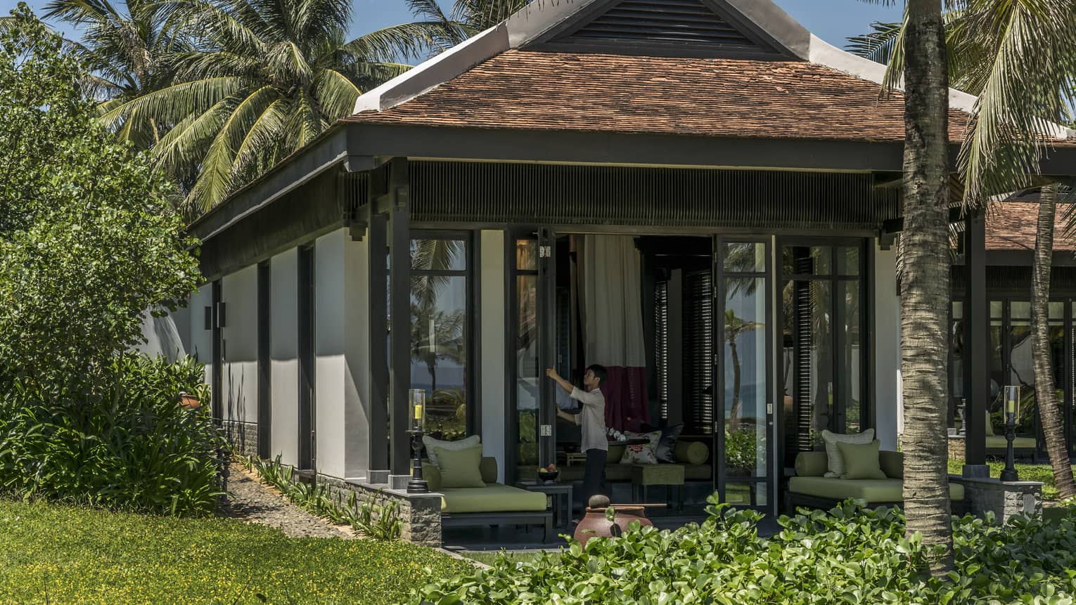 Staff member opens floor-to-ceiling doors of resort villa, framed by palm trees and foliage