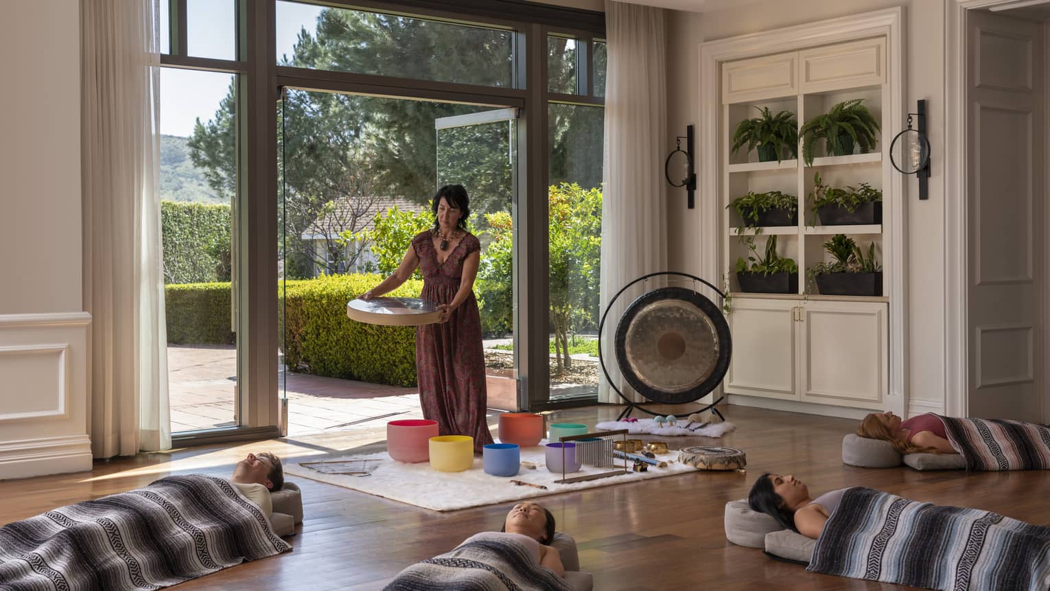A woman standing over colourful buckets with people laying on the floor around her, there is a gong and other instruments.