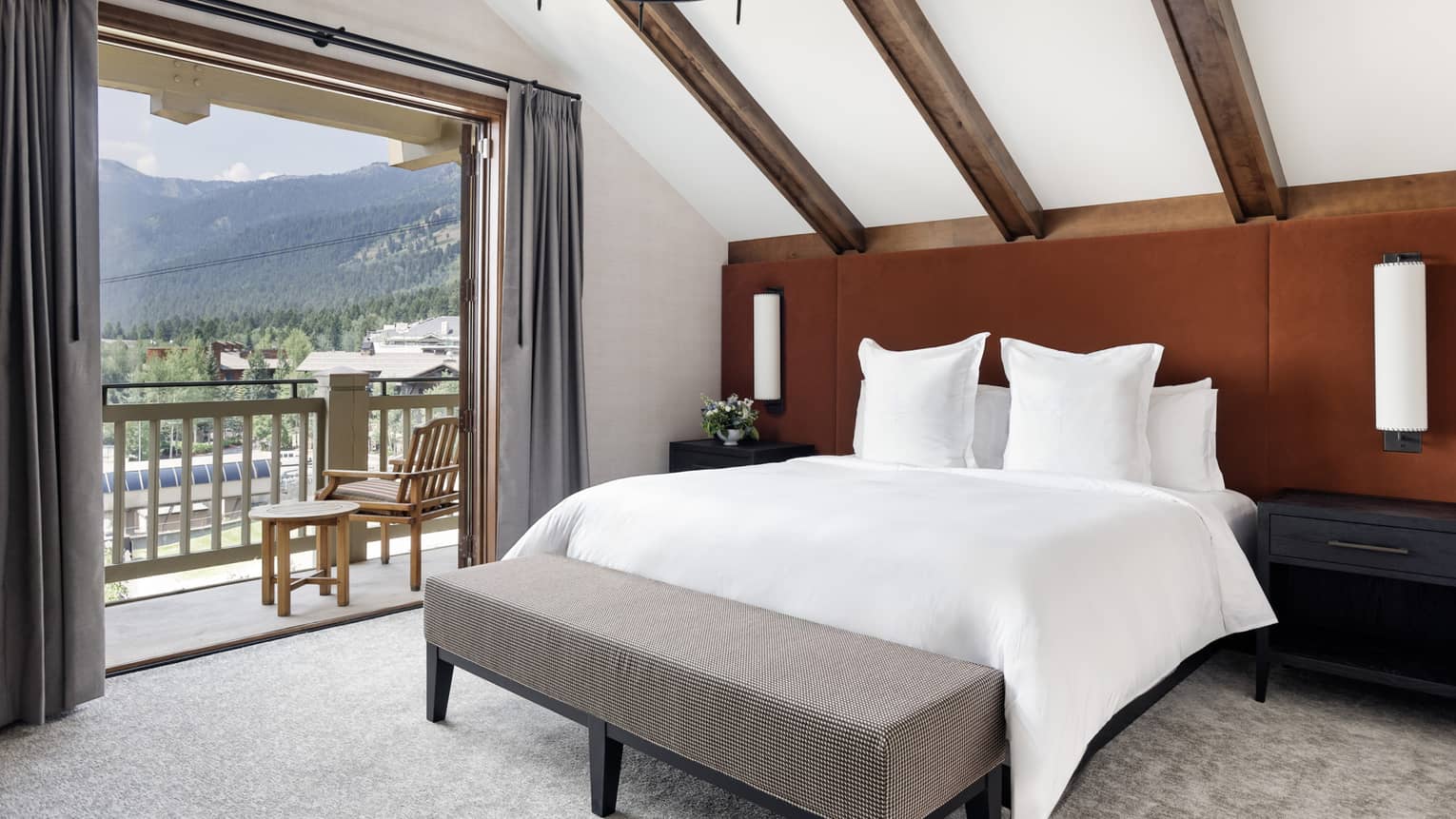 Bedroom with vaulted wood-beam ceiling, rust-coloured headboard, king bed with white linens, grey bench and open balcony with mountain views