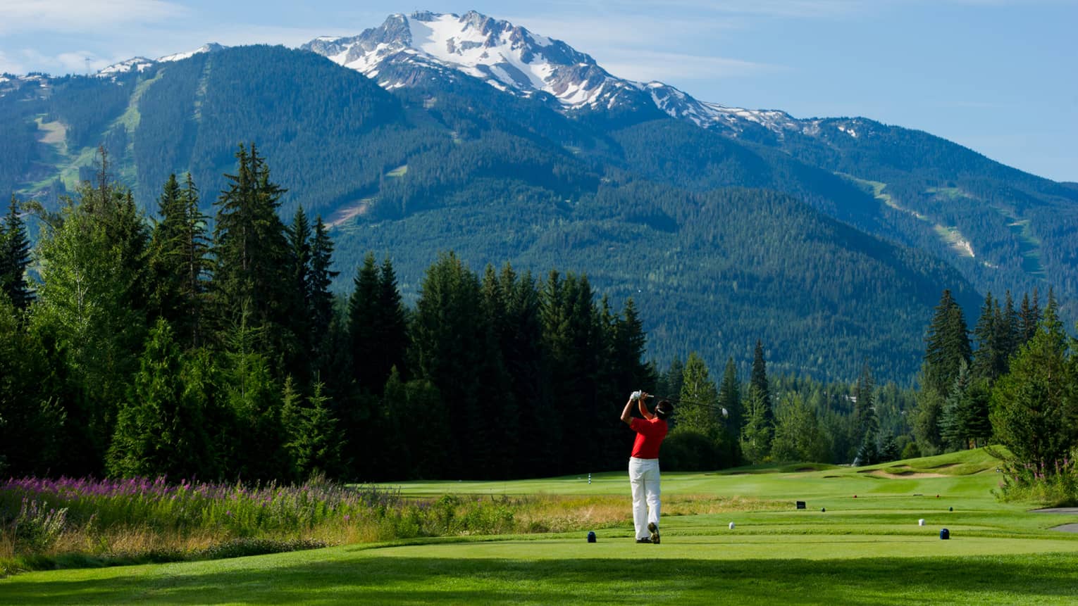 Man wearing red golf shirt swings club on green golf course surrounded by snow-capped mountains
