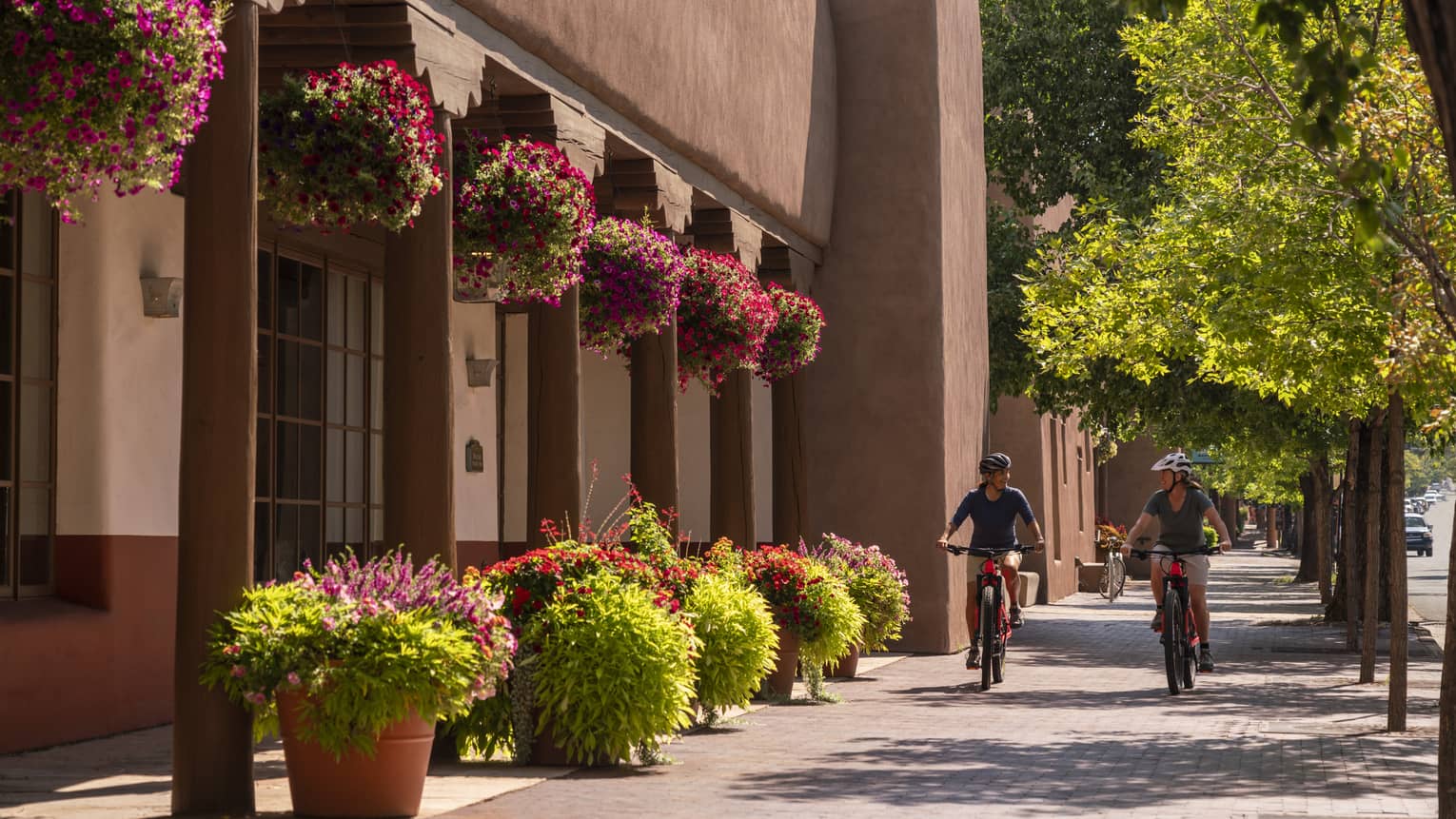 Two people on bikes riding down a small street with a building on one side and small trees on the other.