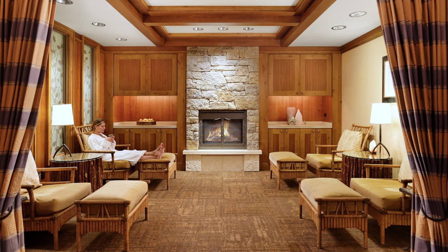 A woman in a spa robe relaxes on a cushioned chair by a lit fireplace