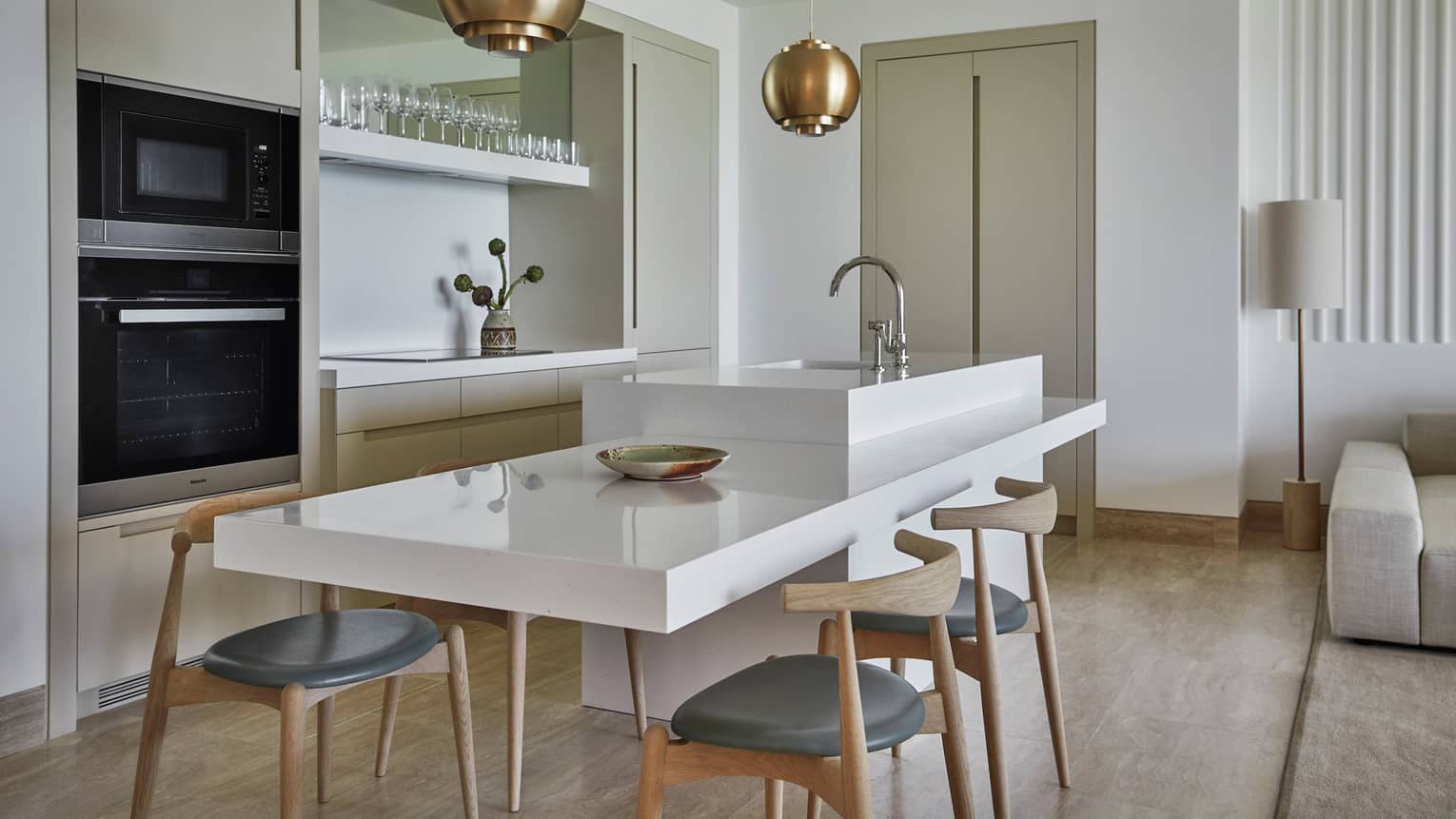 A sleek, modern in-suite kitchen in whites and beiges, featuring a large white serving island with wooden chairs and a sink.