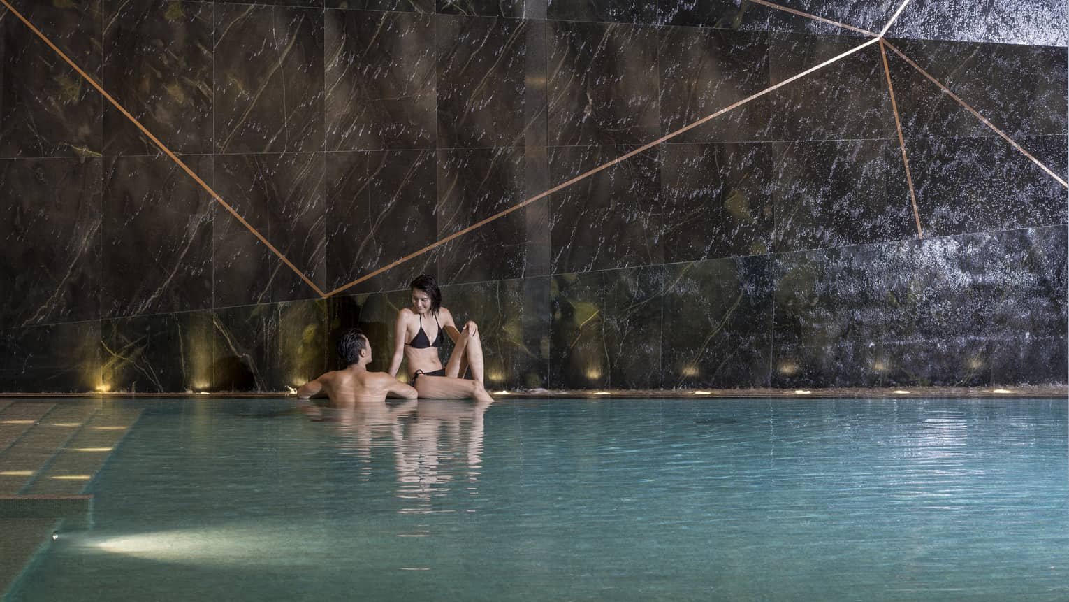 Woman sits on edge of indoor swimming pool, man wades beside her 