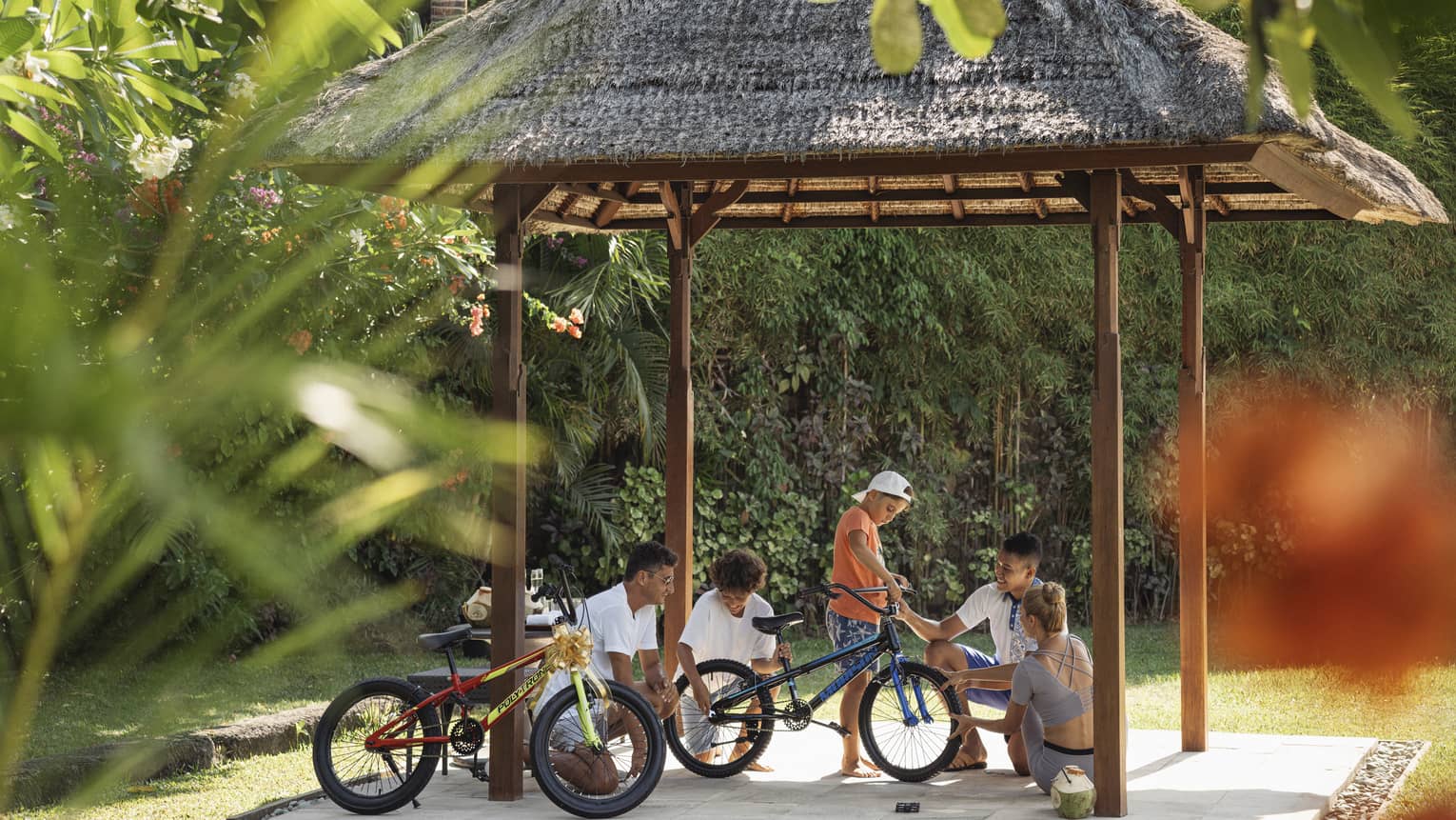 Four seated, smiling adults and a standing child work on a bicycle under a thatched hut surrounded by lush greenery.