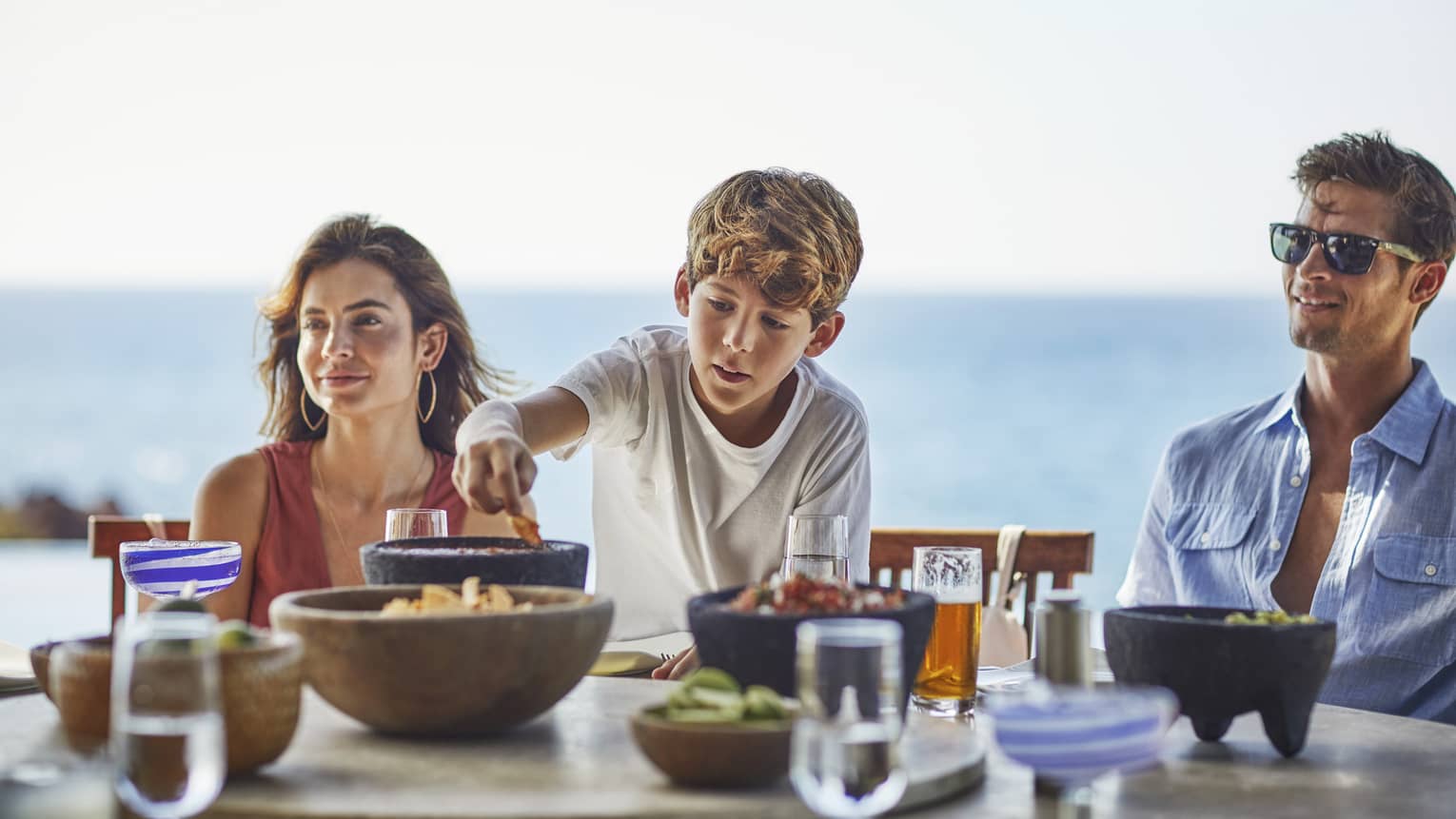 A young boy reaches for a chip bowl on table as a woman and man sit on either side, ocean in the background