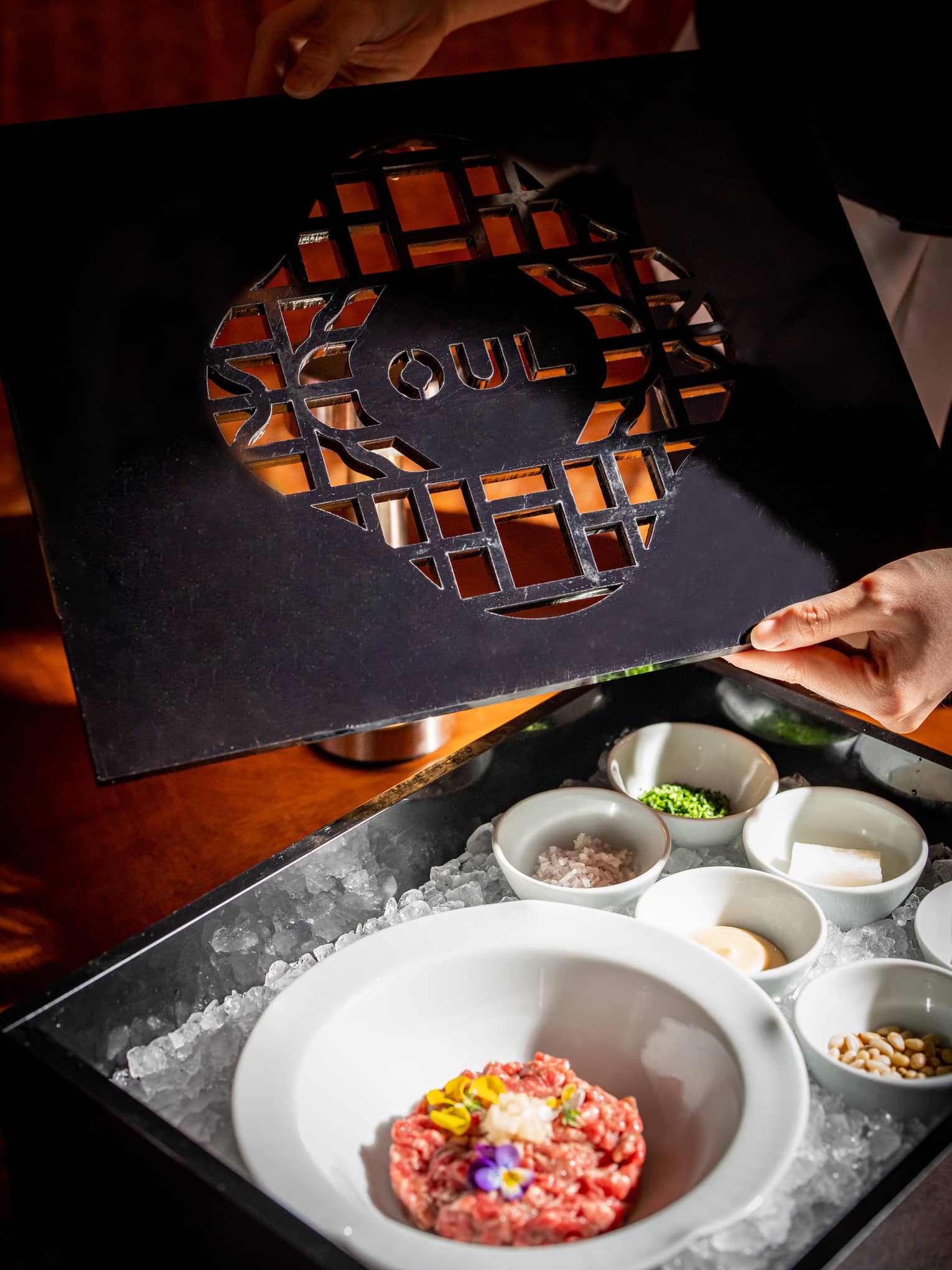 White bowl with beef tartare garnished with flowers, six colourful side dishes, all in a black lacquer ice-filled tray.